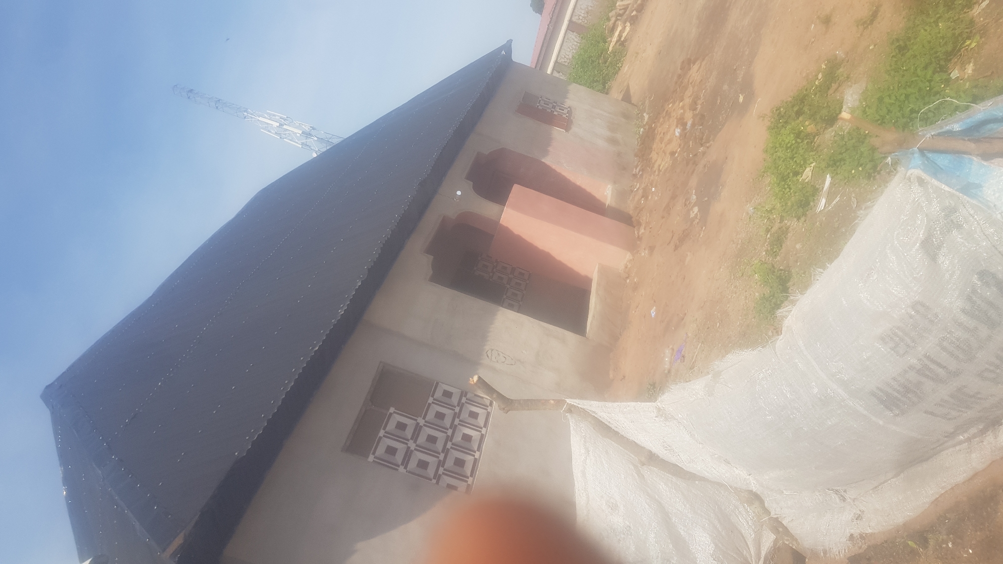 An exterior shot of a single-story building under construction is captured, with a black corrugated metal roof and light-colored walls. The building has two arched doorways on the front facade and two windows with decorative patterned glass. A telecommunications tower is visible in the background, suggesting an urban or semi-urban environment. In the foreground, large white woven sacks are piled up, possibly containing building materials. The ground is unpaved and appears to be dirt, with some sparse vegetation growing. The sky is clear and blue.