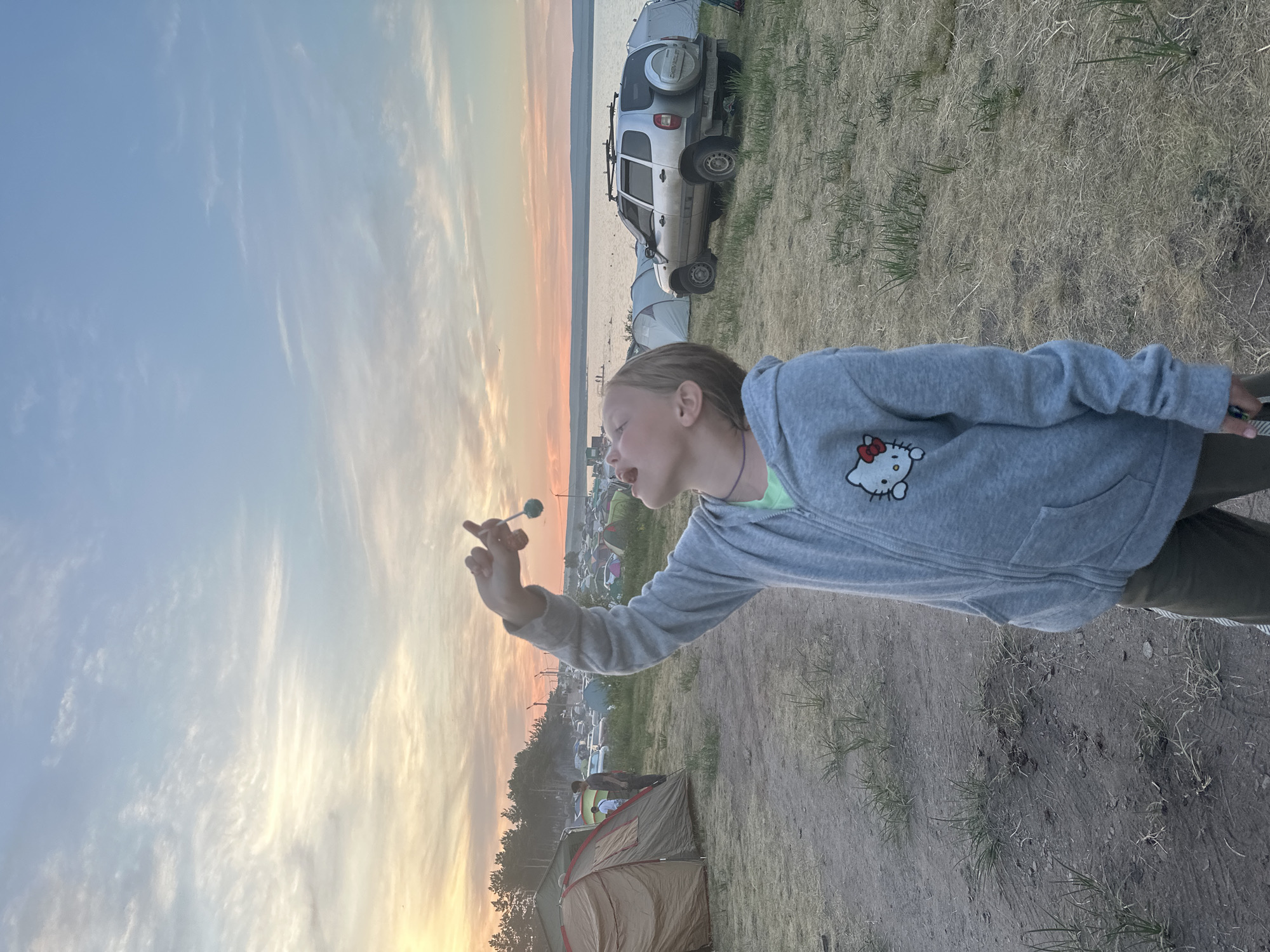 A young person with blonde hair pulled back is standing in a field with dry grass and dirt. They are wearing a gray hooded sweatshirt with a Hello Kitty design and dark pants. The person is looking up and holding a blue, round object on a string to their mouth. In the background, there is a body of water, a shoreline with trees, and a campsite with several tents and a silver SUV. The sky is a soft blend of blue, pink, and orange, suggesting it is either sunrise or sunset. This scene is likely in the city of Zhemchuzhny, Russia, given the presence of the campsite and natural landscape.