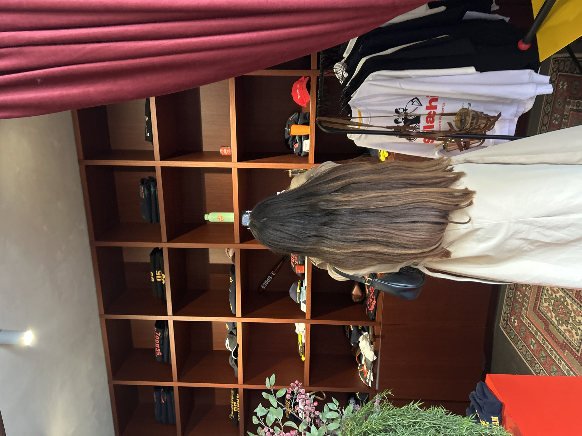 A woman with long, highlighted hair stands in front of a wooden shelving unit in what appears to be a retail space in Yerevan, Armenia. The shelves are filled with items including hats, clothing, and bottles. The woman is wearing a light-colored jacket and a black purse. A burgundy curtain hangs above the shelving unit, and a patterned rug is visible on the floor. A plant is in the foreground.