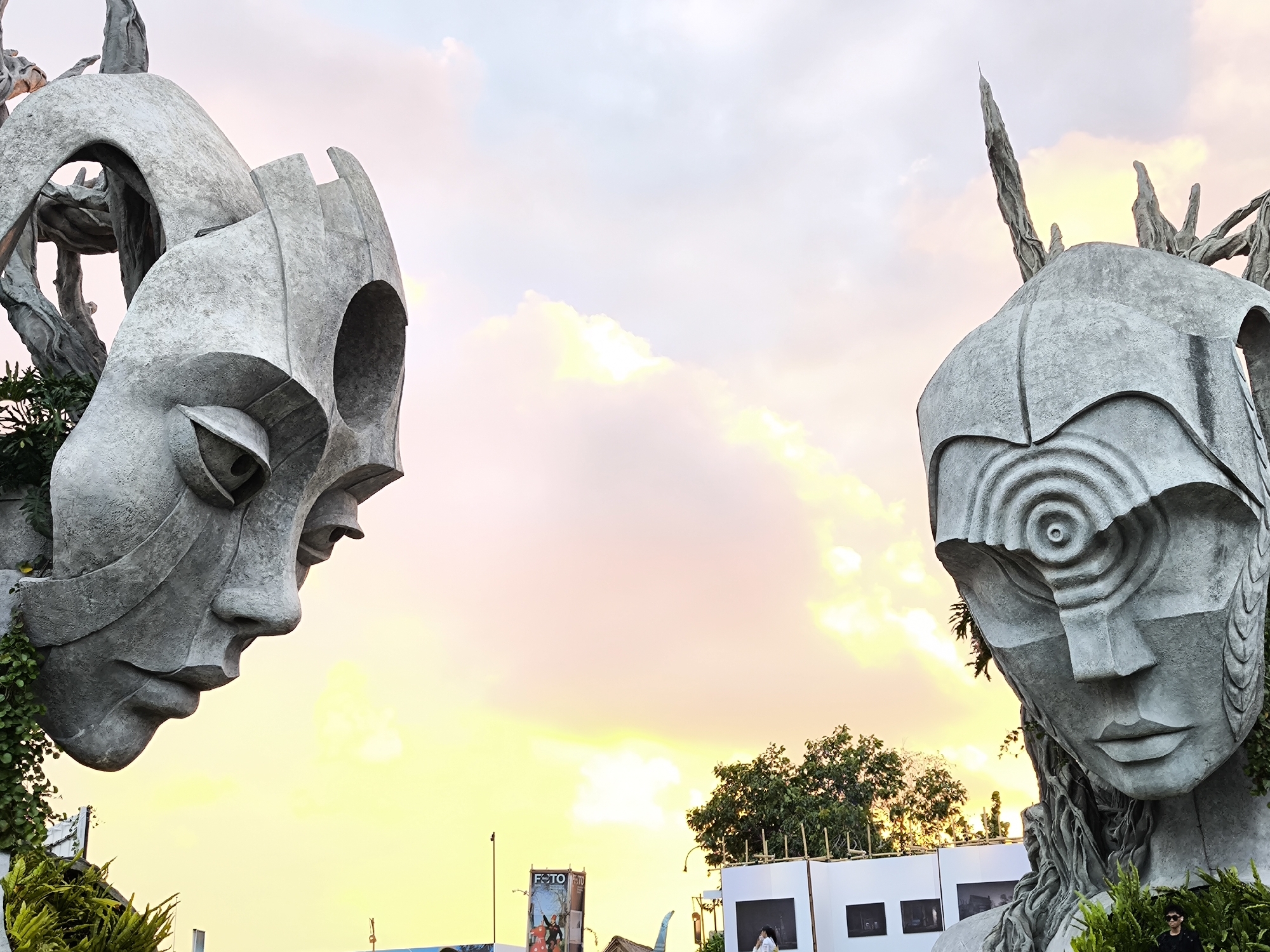 This is a medium shot, eye-level, outdoor photograph of two large, gray, abstract sculptures of human faces. The sculpture on the left is on the left side of the frame, angled towards the right. It features an elongated face with prominent cheekbones, a sharp jawline, and hollowed-out eye sockets. Green vines and foliage are growing around the left side of this sculpture. The sculpture on the right is on the right side of the frame, angled towards the left. It has a rounded forehead with concentric circles, a flat nose, and a defined mouth. Green vines and roots are also visible on this sculpture.
The sculptures are positioned against a backdrop of a sky with soft, warm clouds, illuminated by what appears to be a sunset or sunrise. The sky transitions from a bright yellow near the horizon to a pale pink and then a light blue at the top.
In the lower portion of the frame, there are hints of a city environment. White panels, possibly part of an exhibition or event, are visible behind the sculptures. A billboard with the word "FOTO" is present in the mid-ground. In the bottom right corner, a person wearing sunglasses and a dark shirt is partially visible, standing amidst green foliage. The sculptures appear to be a prominent installation in the city of Kediri, Indonesia, likely part of a public art display or festival.