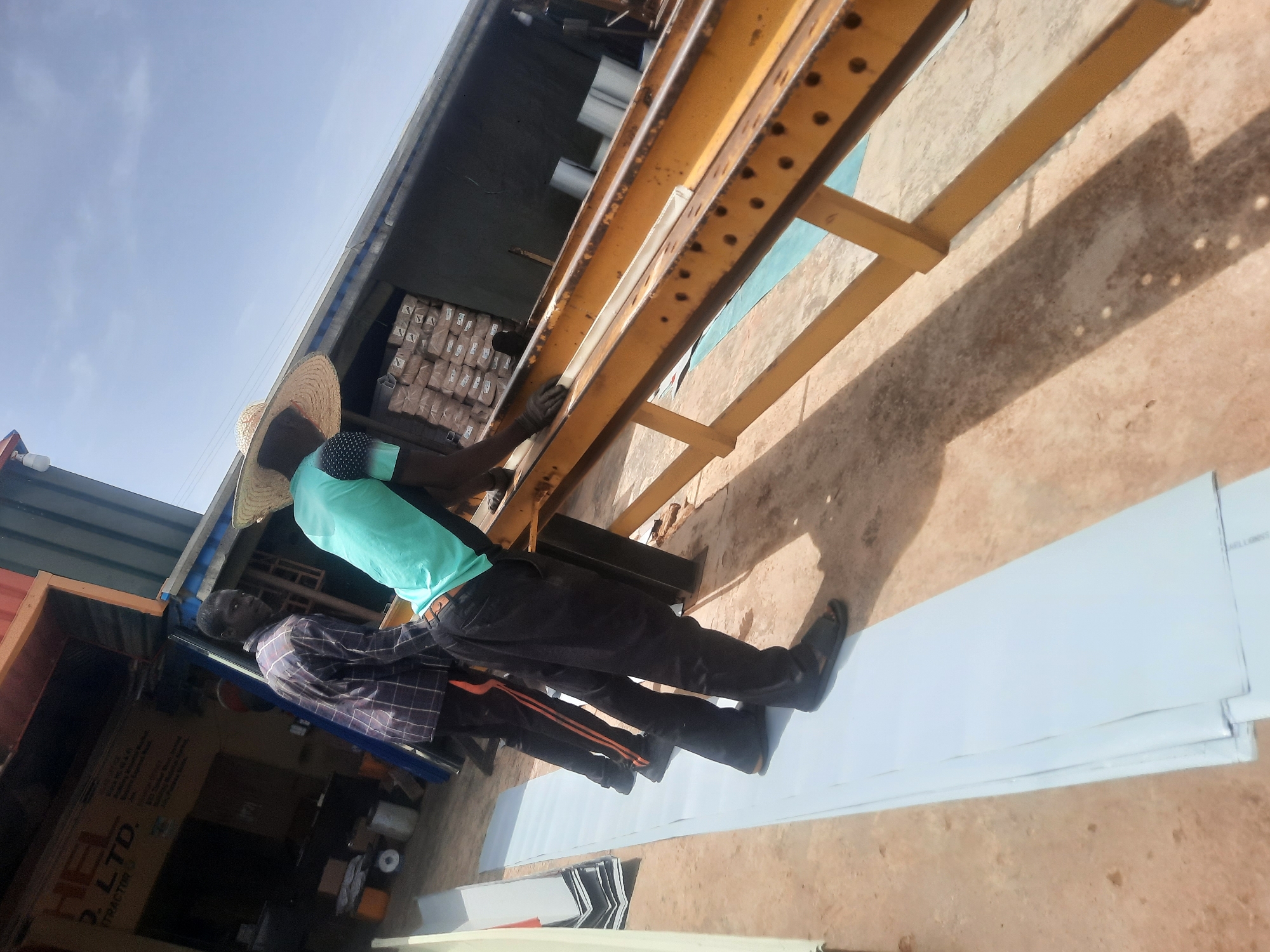 Two adult males are engaged in a construction-related activity in Gura Top, Nigeria. One male, wearing a straw hat and a turquoise t-shirt, is operating a yellow metal press. The other male is dressed in a blue and white plaid shirt with black pants and orange stripe. They are working with sheet metal, with several pieces already laid out on the ground. The scene takes place outdoors, in front of a building. The building has stacks of packaged goods visible under a metal roof, possibly building materials. A sign with "HEL 2. LTD." is partially visible on the left side.