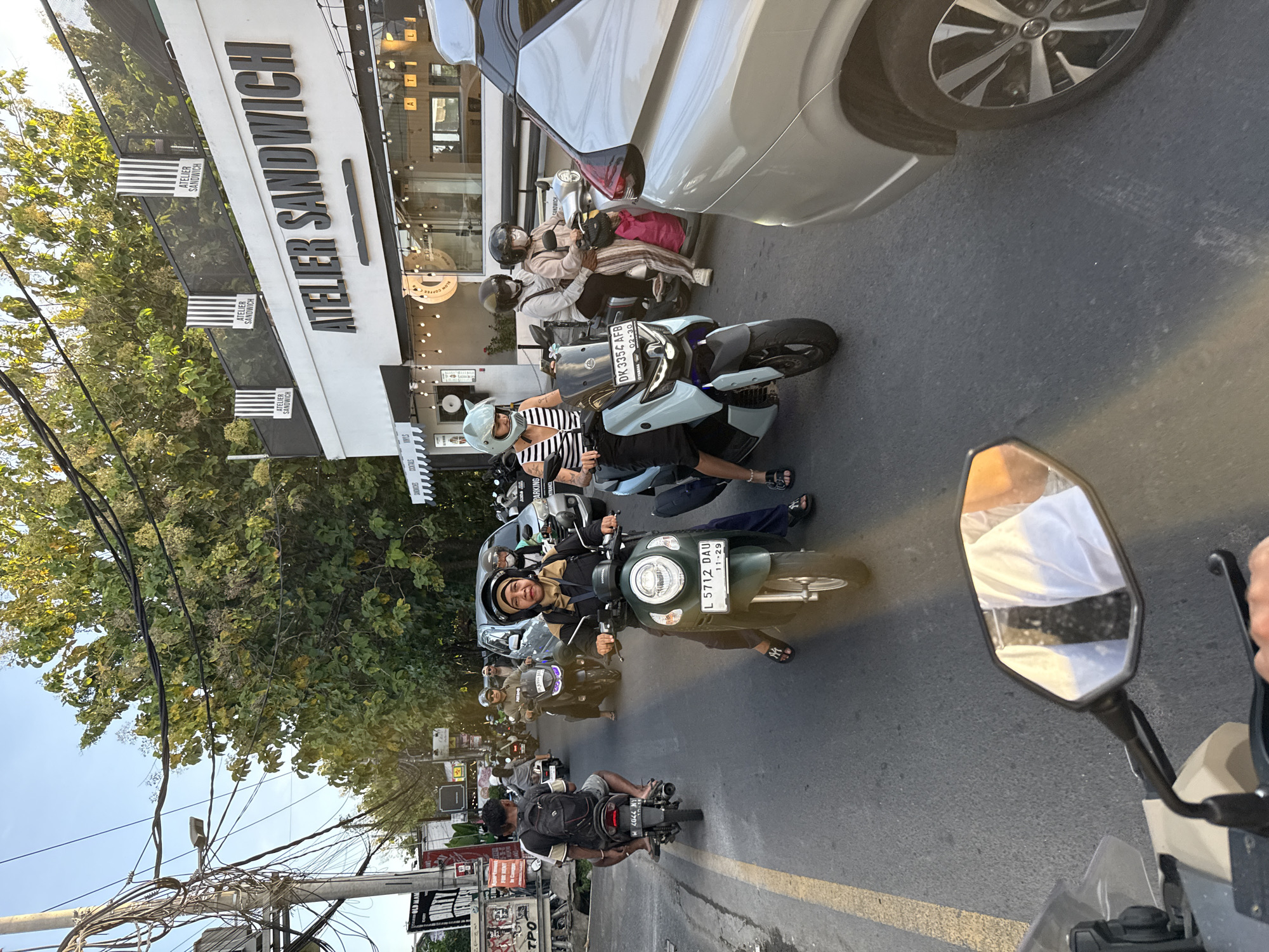 The image captures a bustling street scene in North Kuta, Indonesia, filled with scooters weaving through traffic. The foreground features the side mirror of a scooter, suggesting the photograph was taken from a moving vehicle. Several other scooters are visible, occupied by riders wearing helmets. One female rider is looking directly at the camera, and a female rider behind her is wearing a striped shirt.
In the background, a building stands with the prominent sign "ATELIER SANDWICH," repeating vertically along its facade. The presence of trees, power lines, and additional street signs indicate the location is a busy commercial area. Other riders are visible between the scooters, and a silver car is partially in view on the right side of the frame.