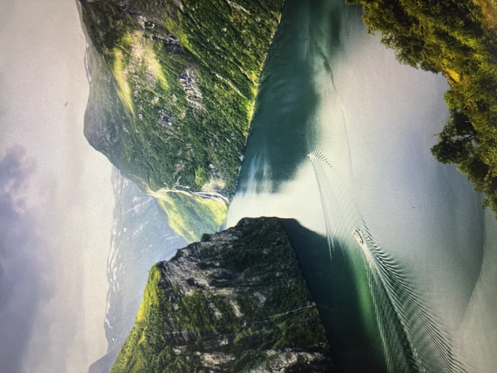The image, captured in Asenovgrad, Bulgaria, displays a majestic natural landscape oriented sideways. It features a narrow, deep green body of water, strongly resembling a fjord, flanked by towering, steep mountains. These mountains are richly covered in vibrant green vegetation, suggesting lush forests or dense shrubbery, with some rock faces visible. On the calm surface of the water, a small boat is visible, actively moving and leaving a long, distinct V-shaped wake that stretches behind it.
The sky in the background is partly cloudy, but the scene is well-illuminated, indicating daytime. The lighting highlights the textures of the mountains and creates reflections on the water. The overall impression is one of dramatic natural beauty and serene activity. The provided crops reveal a prominent pixelated grid, indicating that the original image is likely a photograph of a digital screen displaying this scenic view, rather than a direct photograph of the landscape itself. No visible text is present in the image.