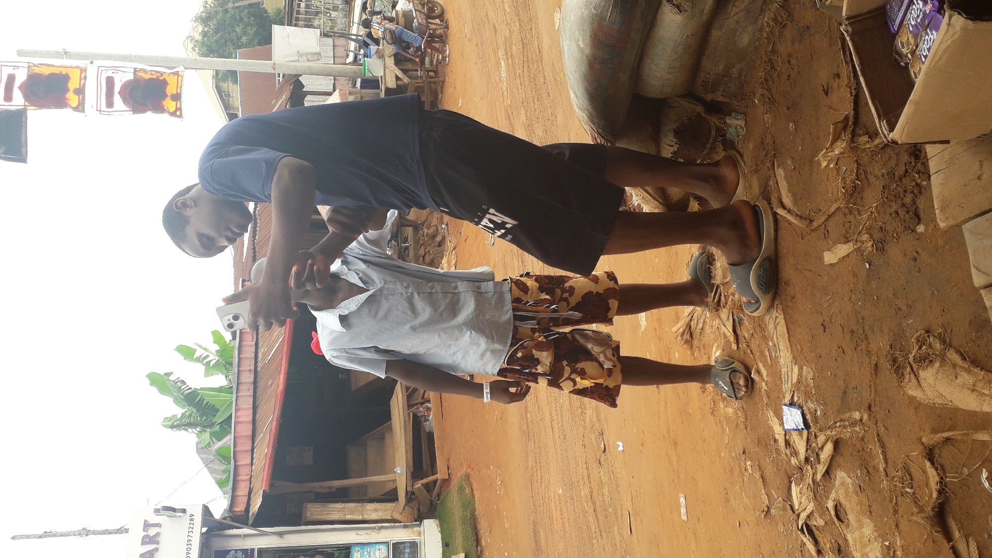 Two young men stand on a dirt road in what appears to be Nkpor, Nigeria. The man in the foreground wears a light-colored button-down shirt and camouflage shorts, while the man behind him wears a dark blue shirt and black shorts. The foreground man holds a smartphone, possibly taking a picture or video of the man behind him. They both wear sandals. The surrounding environment includes buildings, a store with signage, and various objects like bags and cardboard boxes. The ground is dirt-covered, suggesting a market or roadside area. Other people are present in the background.