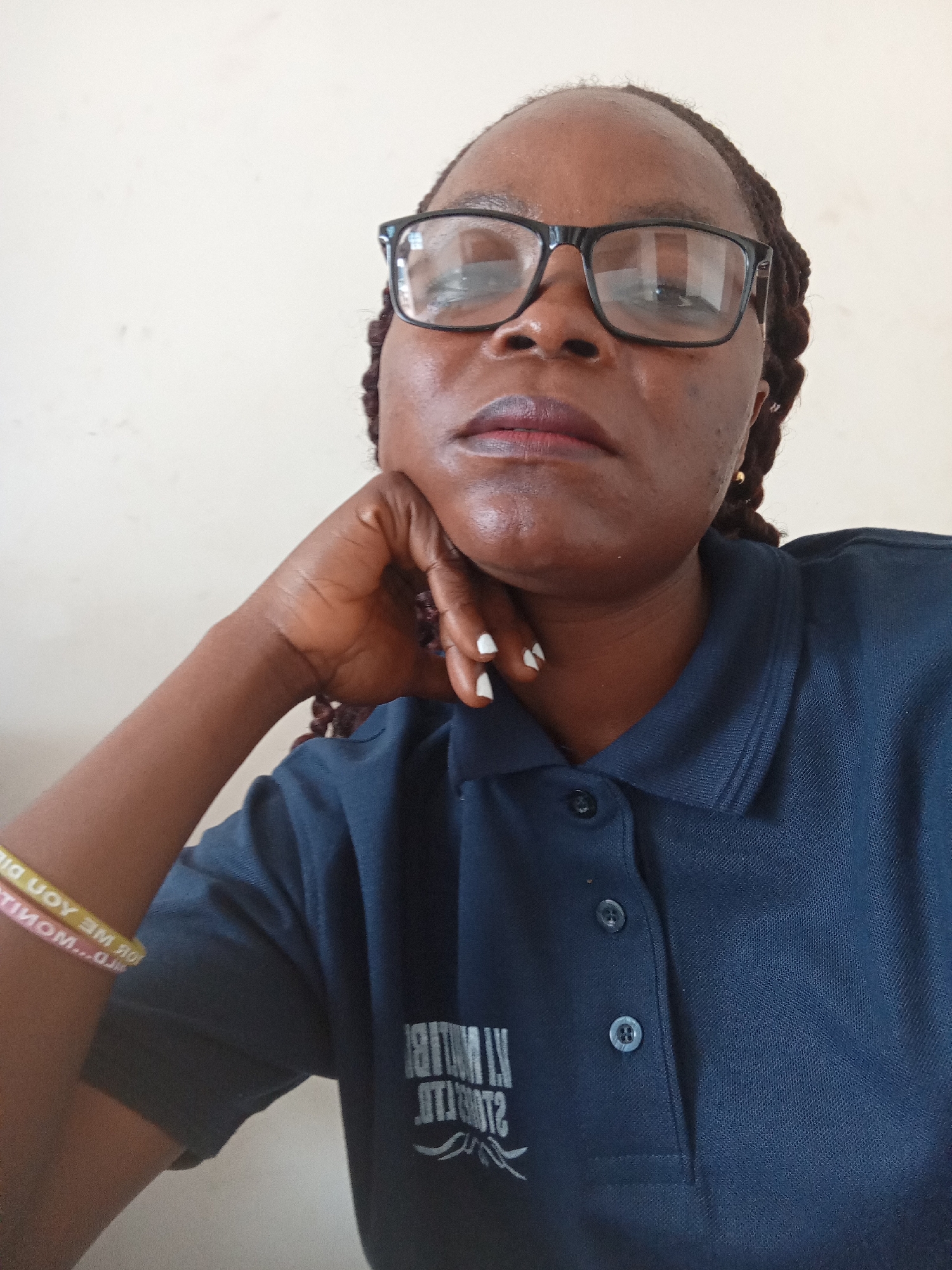 A medium shot shows a Black woman wearing glasses and a dark blue polo shirt. She is resting her chin on her hand, with her elbow on her arm. Her braids are visible, and she is wearing small gold earrings. A yellow and pink bracelet is on her left wrist. The polo shirt has a logo with visible text on the left chest. The background is a plain, light-colored wall, suggesting an indoor setting. The lighting appears to be natural, possibly from a window. The woman's expression is neutral, and her gaze is directed slightly upward. The overall impression is a casual, candid portrait. Text on the bracelet reads "FOR ME YOU DID" and "PLATINUM... GLOW". The logo on the shirt is mostly obscured, but some white text and a graphic element are visible.