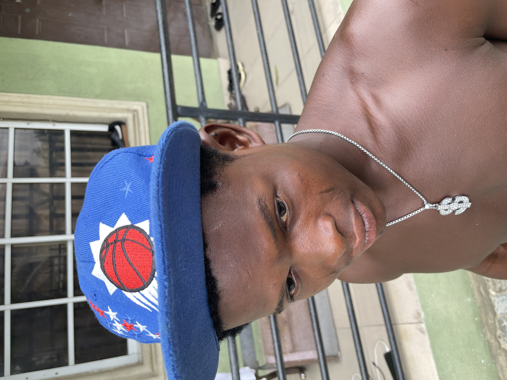A young man with dark skin and a silver necklace featuring a pendant is wearing a blue baseball cap with a red basketball emblem and white and red stars. He is positioned in front of a barred gate, with a green wall and a window visible in the background. The location is implied to be Ado, Nigeria.