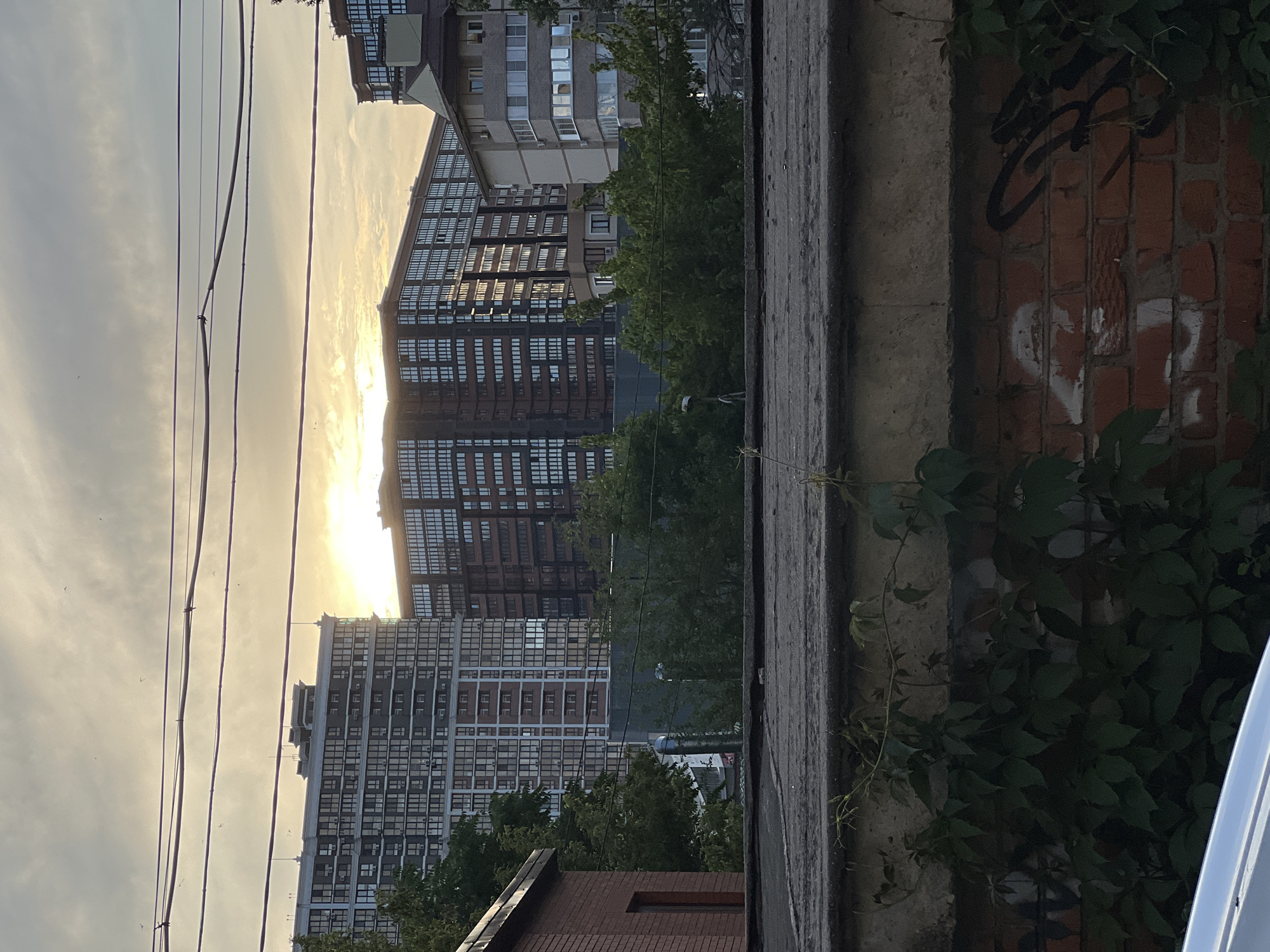This media file depicts an urban scene in Krasnodar, Russia, likely during the late afternoon or early evening due to the low angle of the sun. The setting appears to be a residential area with several tall apartment buildings visible.
The foreground features a textured concrete surface, possibly a wall or ledge, with some green vines growing on it. Adjacent to this is a brick wall with graffiti, including what appears to be stylized lettering and a white heart shape.
In the mid-ground and background, multiple apartment buildings dominate the view. These buildings are characterized by numerous windows, suggesting high occupancy. The architecture suggests a post-Soviet era construction style. Trees and foliage are interspersed among the buildings, indicating a degree of urban greenery.
The sky is overcast with clouds, but the sun is breaking through, casting a warm, golden light on the scene and creating reflections on the windows of the buildings. A series of power lines run horizontally across the frame, adding to the urban feel.
The overall mood is one of everyday urban life, with the setting sun lending a slightly tranquil or contemplative atmosphere. No people are directly visible in the frame, but their presence is implied by the residential setting. No discernible text beyond the graffiti is visible.