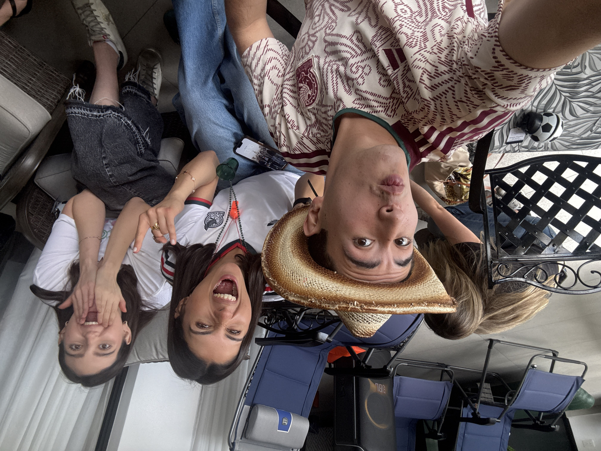 In San Bernardino Tlaxcalancingo, Mexico, four individuals are captured in a candid selfie. Two women are seated on a sofa in the foreground, their expressions animated with excitement, with one woman in the center with her mouth wide open and the other to her right covering her mouth with her hands. Another woman is partially visible behind them, hunched over with her long blonde hair obscuring her face. A man in a cowboy hat and a patterned shirt is in the center, making a kissing face towards the camera. In the background, several blue lounge chairs are arranged around the outdoor seating area. A small soccer ball rests on a patterned cushion on the floor. The lighting suggests it is daytime, and the setting appears to be a residential patio or outdoor entertainment space.