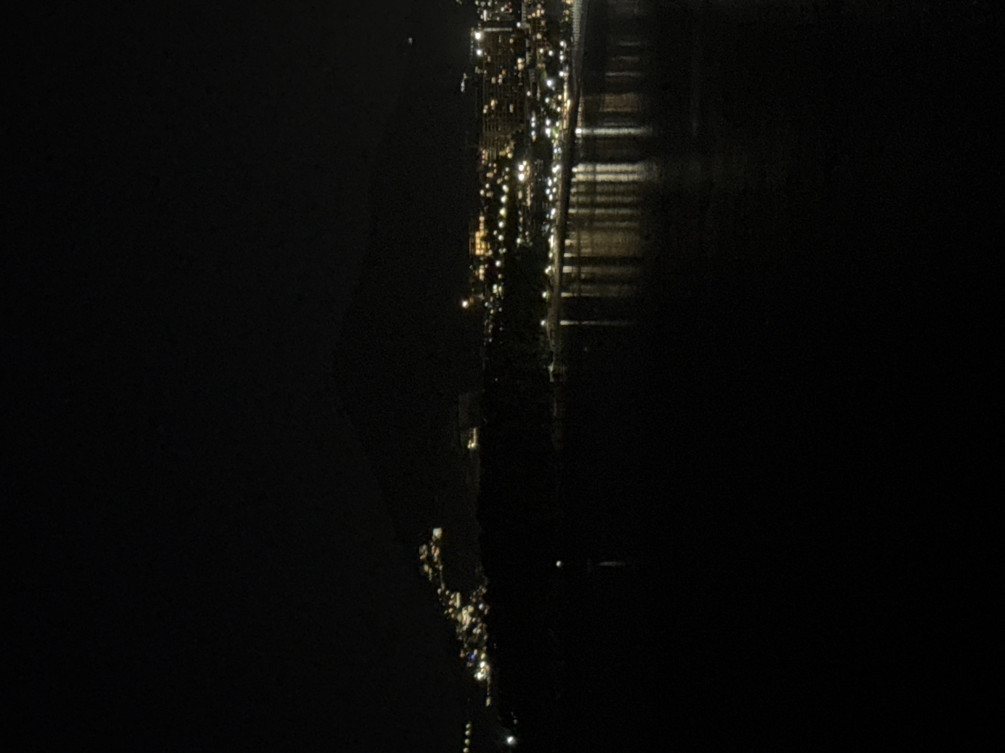 An aerial nighttime view depicts the coastline of Rafailovići, Montenegro, characterized by a linear stretch of illuminated buildings and streetlights along the shore, adjacent to a body of water. The lights create reflections on the water surface. To the left, land mass fades into darkness, and on the right, the water extends into blackness.