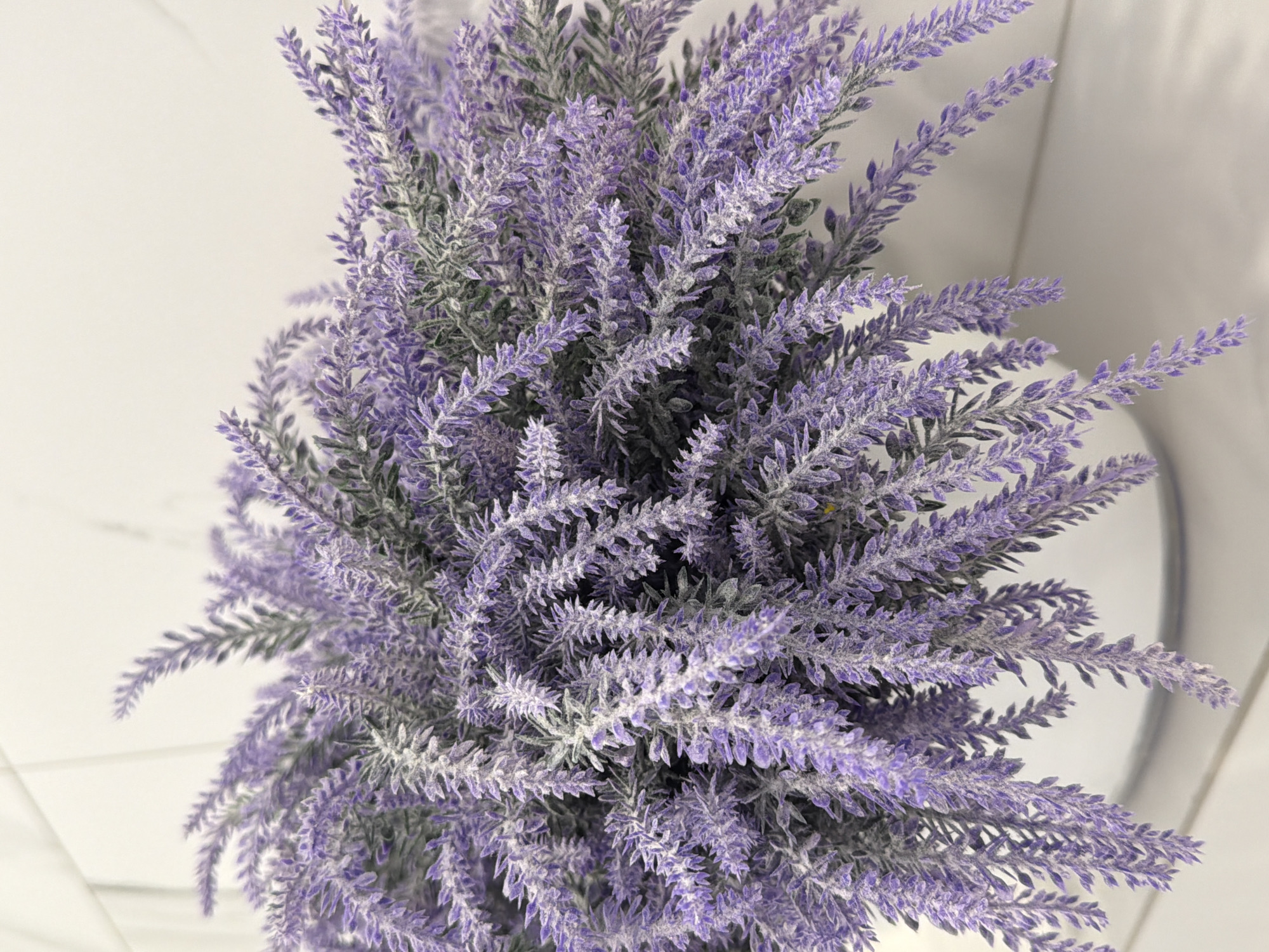 A close-up, overhead shot of artificial lavender in a white pot. The lavender is a deep purple color with a dusty white coating. The background is a soft, blurred white, suggesting a clean and minimalist setting. There are no people or animals in the image. The scene is a still life, with no action taking place. The lighting is soft and even, highlighting the texture of the lavender. The overall impression is one of calm and serenity. The location in the city of Kazan, Russia is not discernible from the image.
