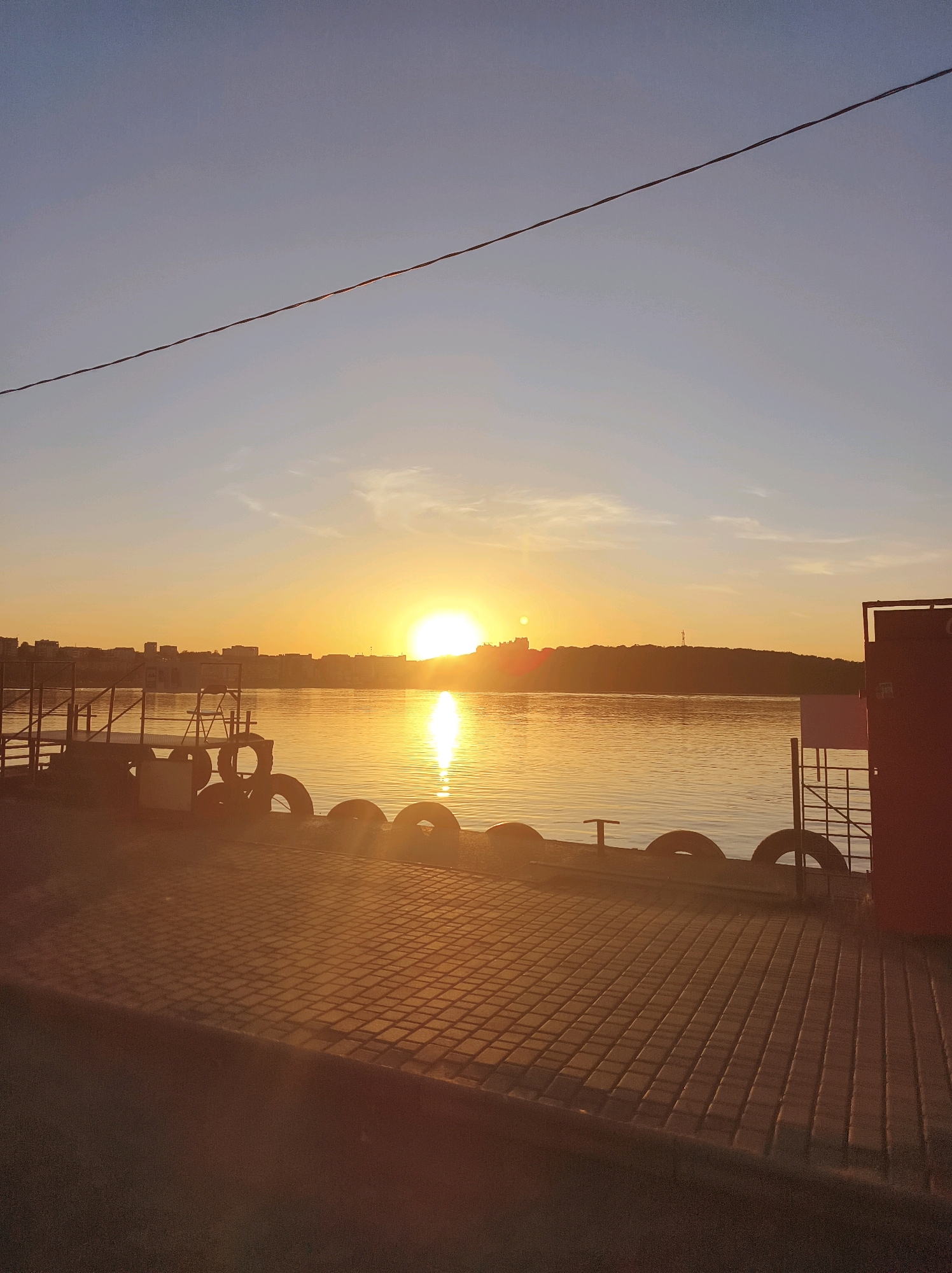 The image captures a serene sunset over a body of water, likely the Ternopil Reservoir, in Ternopil, Ukraine. In the foreground, a paved area with a tiled surface leads to a dock or pier. Several old tires are stacked along the edge of the dock, serving as fenders. A metal railing and stairs are visible on the dock. To the right, a red container or structure stands prominently. The water reflects the warm golden hues of the setting sun, which is positioned low on the horizon, casting a bright glow. Across the water, a city skyline with buildings and a distant tower is silhouetted against the sky. A thin wire stretches across the upper portion of the frame. The overall atmosphere is peaceful and tranquil, characteristic of a lakeside evening.