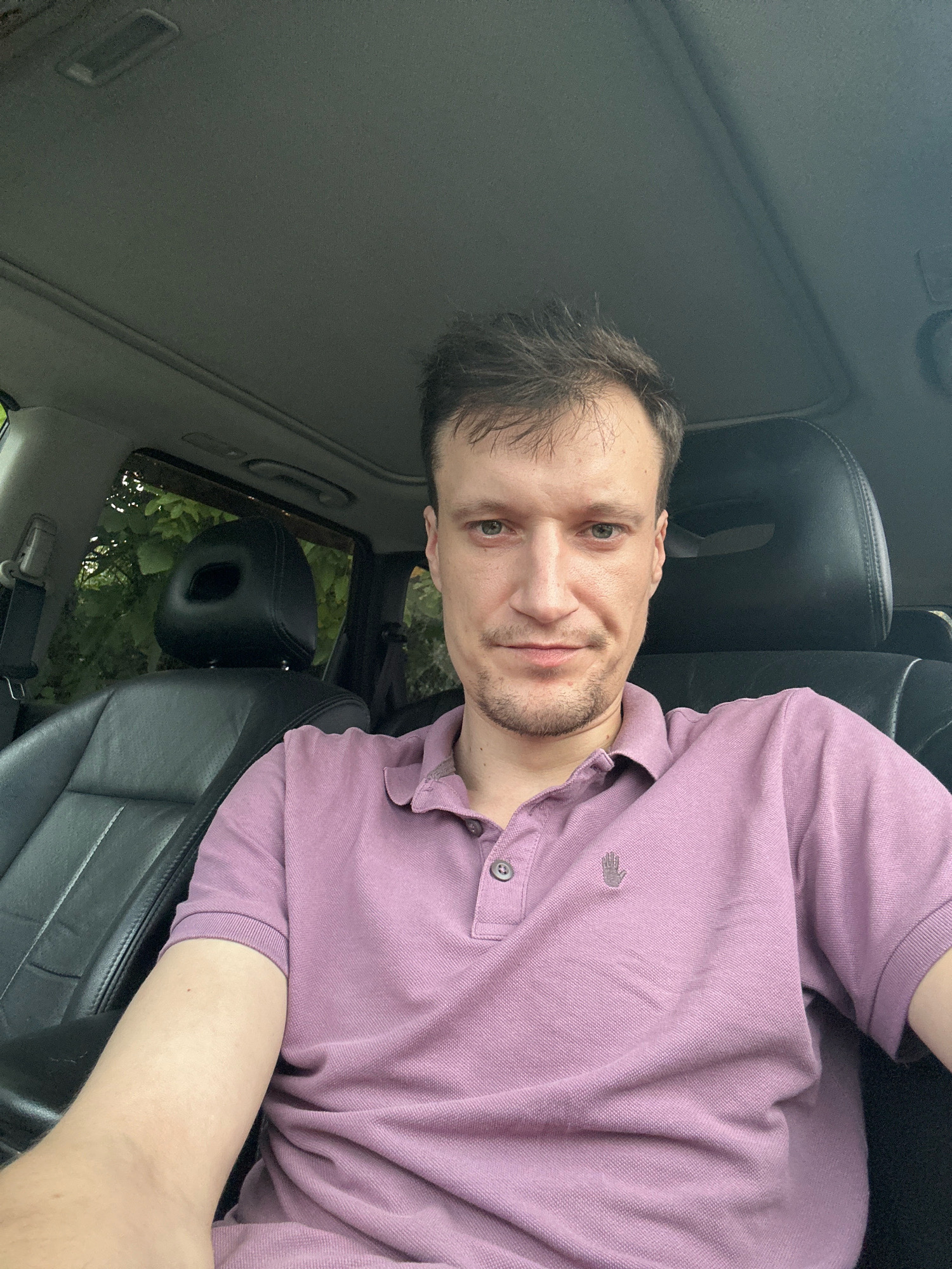 In the city of Shatava, Ukraine, a man with light skin, short brown hair, and light green eyes is captured in a selfie taken from inside a car. He is wearing a purple polo shirt with a small embroidered logo on the left side of his chest. The man is seated in the driver's seat, with the car's black leather interior visible, including the headrest and part of the passenger seat. The background shows lush green foliage through the car windows, suggesting an outdoor, possibly wooded, environment. The car appears to have a sunroof, visible in the upper portion of the image. The man's expression is neutral, and he is looking directly at the camera.