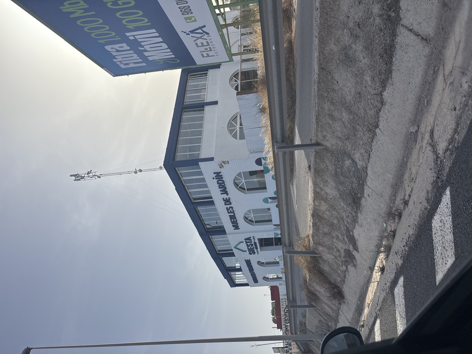 The image depicts a commercial building located in Urbanización Cabo Roig, Spain. The building is a two-story structure with a white facade and a dark blue roofline. The ground floor features arched windows and the upper floor has rectangular windows. Prominently displayed on the building are signs that read "MUEBLES DE JARDÍN" (Garden Furniture) and "SIESTA GROUP." To the right of the building, a large billboard advertises "Fibra 1.000 Mb" and "Móvil 100 GB" along with "FLEXA NETWORKS." In the background, a cell tower is visible against a clear sky. The building is situated adjacent to a road with a metal guardrail in the foreground. A drainage ditch runs alongside the road, with dry vegetation and dirt. The perspective suggests the photo was taken from a moving vehicle.