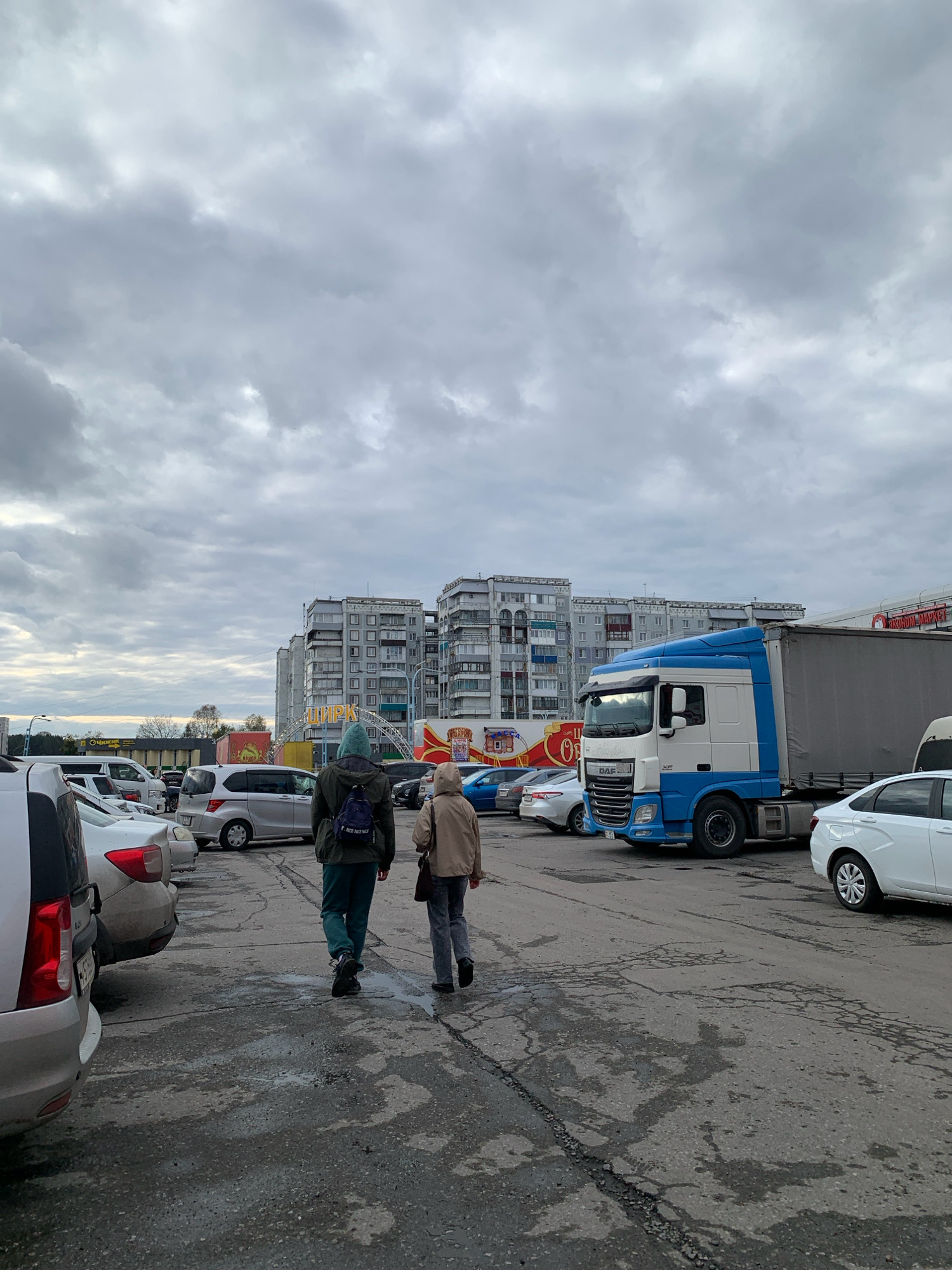 Two individuals are walking across a paved area in front of a row of apartment buildings. The person in front is wearing a dark green hooded jacket with a blue backpack and dark pants. The person behind is wearing a light brown hooded jacket and dark pants. They are walking away from the camera and towards the apartment buildings. In the background, there are several parked cars, including a white truck with a blue cab, and a large white semi-trailer truck. A large sign with Cyrillic lettering, reading "ЦИРК" (CIRCUS), is visible above the parked cars. The sky is overcast with gray clouds. The ground is wet and appears to be cracked asphalt. The scene is likely a parking lot or an open area near residential buildings and a commercial establishment.