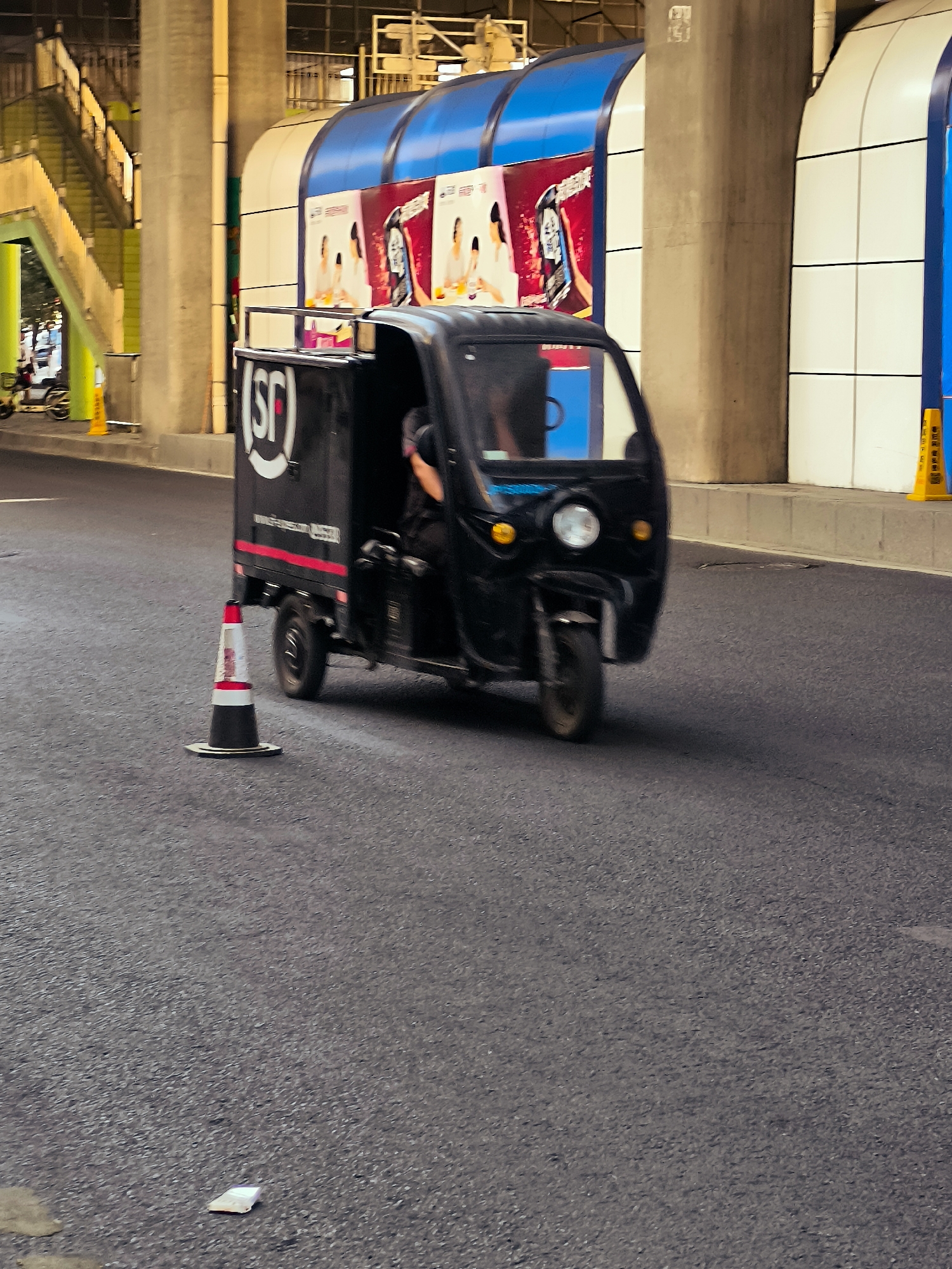 A black three-wheeled cargo vehicle with an "SF" logo on its side is in motion on an asphalt road. The vehicle has a driver partially visible within. A red and white traffic cone with a reflective band is positioned on the road to the left of the vehicle. In the background, a modern elevated structure with blue and white panels and advertisements is visible. Yellow traffic cones are also present in the background. The scene appears to be an urban environment in Ürümqi, China, given the distinctive architecture and advertisements that are characteristic of the region.