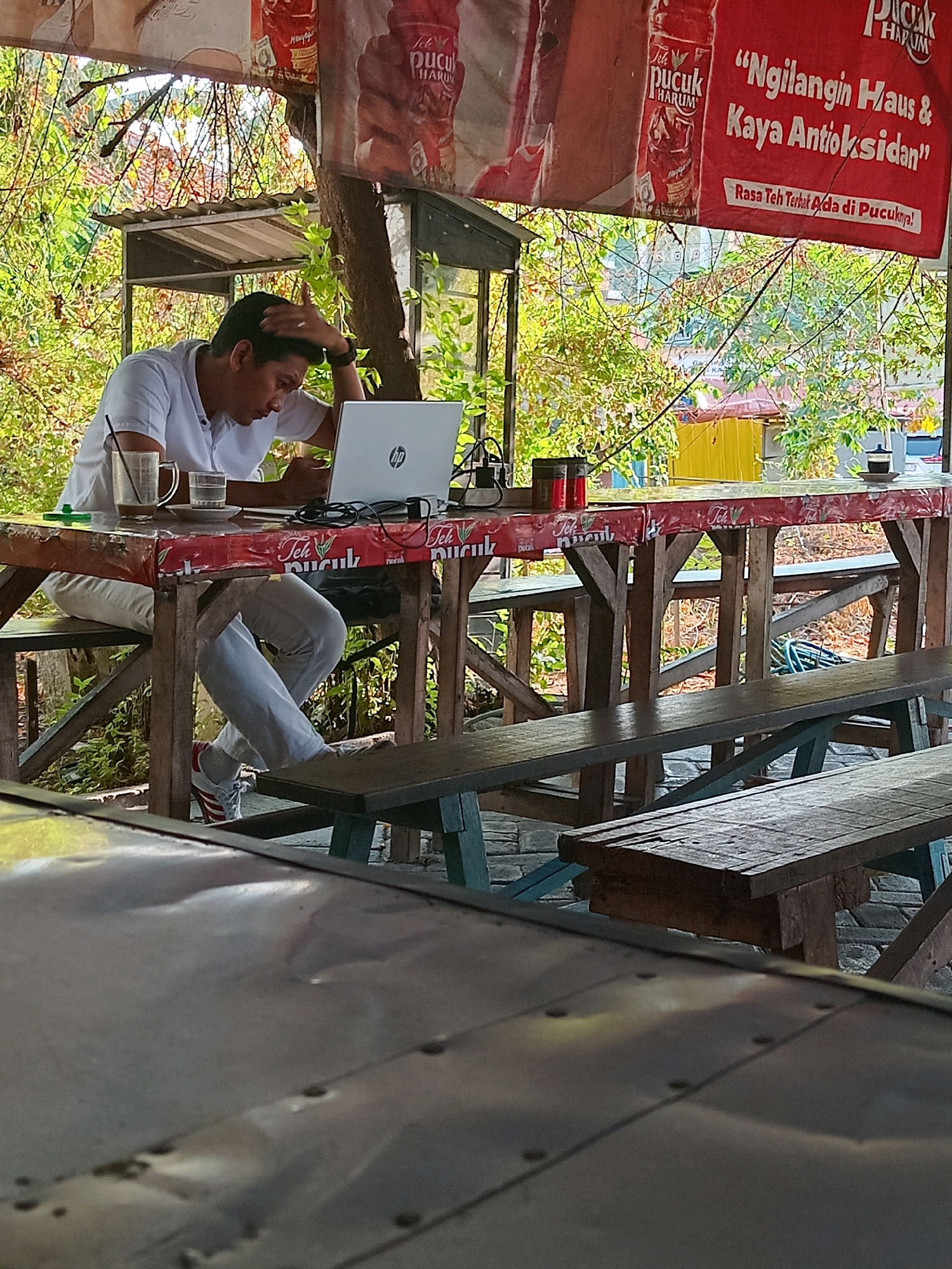In Kembangan, Indonesia, a man in a white polo shirt and light-colored pants sits at a wooden table covered with a red and white patterned tablecloth, working on a silver HP laptop. He has his right hand on his head, appearing to be concentrating or stressed. On the table, there are two glasses, one with a straw and a dark liquid, and another with water, along with a cup and saucer. Behind him, a banner advertising "Teh Pucuk Harum" hangs, featuring Indonesian text. The scene is outdoors, with lush green foliage visible in the background. Wooden benches are positioned around the table.