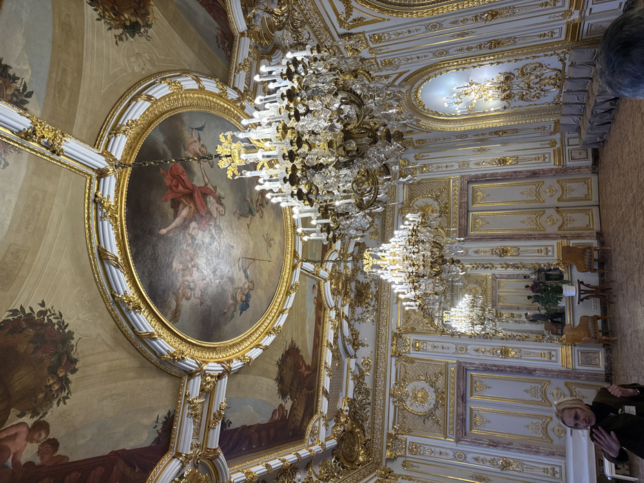 The image depicts a grand hall within the Winter Palace, part of the State Hermitage Museum, in Saint Petersburg, Russia. The room exhibits an opulent, Baroque-revival style characterized by extensive gilding, frescoed ceilings, and crystal chandeliers.
The high ceiling features a large central oval fresco depicting a figure in red drapery amidst clouds and cherubic figures, framed by elaborate gilded moldings. Surrounding this central painting are smaller rectangular and octagonal ceiling panels, also adorned with frescoes of putti, garlands, and allegorical scenes, all set within gilded decorative borders. The ceiling structure also incorporates white and gold cornices and intricate relief work.
The walls are predominantly light-colored, likely off-white or cream, heavily decorated with gilded stucco work, carved panels, and classical motifs including pilasters and floral scrollwork. Several large arched openings are visible; some contain closed, gilded double doors, while others hold tall mirrors reflecting portions of the opposite wall and room. Wall-mounted gilded sconces with multiple candle-shaped electric bulbs are present on the right wall.
The room is illuminated by three prominent, multi-tiered crystal and gilt-bronze chandeliers, each equipped with numerous lit electric candle bulbs. Additional light sources come from the wall sconces. The floor features a patterned parquet design in shades of brown.
In the left-midground, two ornate wooden chairs with upholstered seats flank a small, round wooden table topped with a potted green plant. Against the right wall, a row of approximately nine identical chairs, each draped in a light grey or silver fabric cover, is neatly aligned.
In the left-foreground, a woman is partially visible from the chest up. She has short, light-colored hair, partially covered by a light headwrap or scarf, and wears a dark top with a visible lanyard and ID badge. Her right hand is raised, palm open, in an active gesturing posture, and her head is turned slightly to her left. In the lower right foreground, the top of a person's head with short grey hair is visible. In the background, reflected in a large wall mirror, three indistinct individuals appear to be standing within the hall.