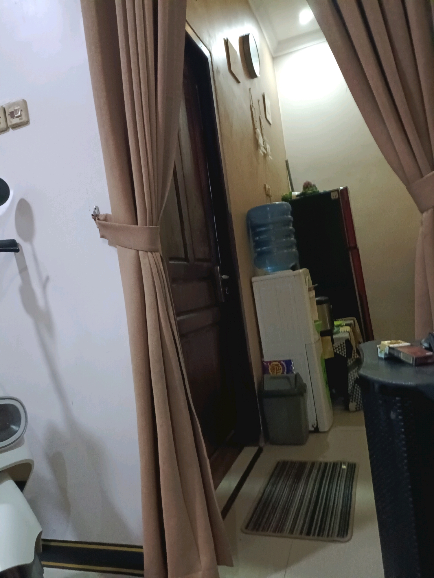 The image depicts an indoor scene, likely a room in a residential or commercial building in Rancang Kencono, Indonesia. The foreground is dominated by a large, light brown curtain that is partially drawn, obscuring a wooden door. To the left of the curtain, a white wall features two light switches. Further to the left, a white appliance with a metallic circular component is partially visible, possibly a fan or a part of a scooter. The middle ground reveals a water dispenser with a large blue water bottle on top, situated next to a white appliance that could be a refrigerator or another cooler. Behind these, a large, dark refrigerator stands against a beige-colored wall. The floor is tiled with a striped rug placed in front of the water dispenser. In the background, a clock and a mirror are mounted on the wall. The overall lighting suggests it is daytime, with light emanating from an unseen source and reflecting off the ceiling.