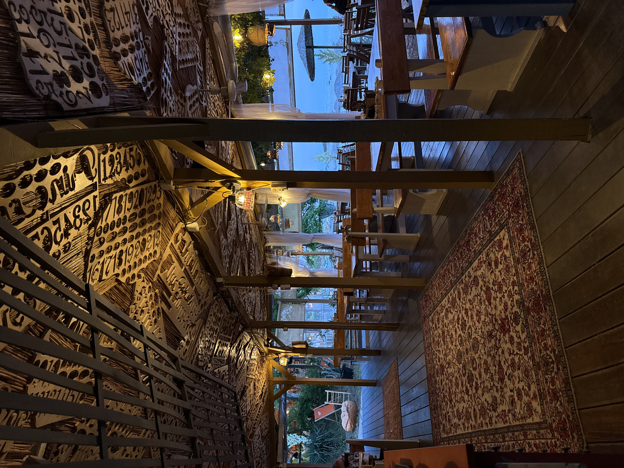 This image depicts an outdoor dining area at a restaurant in Tsarevo, Bulgaria. The scene is set at dusk, with warm artificial lighting illuminating the space. The restaurant features a wooden deck floor and a rustic roof constructed from what appears to be corrugated metal or wood panels, decorated with a collage of various numbers, letters, and circular shapes. Several wooden tables and benches are arranged for seating.
In the foreground, two traditional Persian-style rugs are laid out on the wooden deck. To the left, a red lounge chair and a large, round, orange beanbag are visible in a more relaxed seating area. In the background, beyond the dining tables, the sea is visible with a thatched umbrella providing shade on the beach. White curtains hang from the structure, adding a soft, airy feel. Two ceiling fans are also mounted on the underside of the roof. One person is partially visible sitting at a table in the far right of the image. The overall atmosphere suggests a casual, open-air dining experience by the sea.