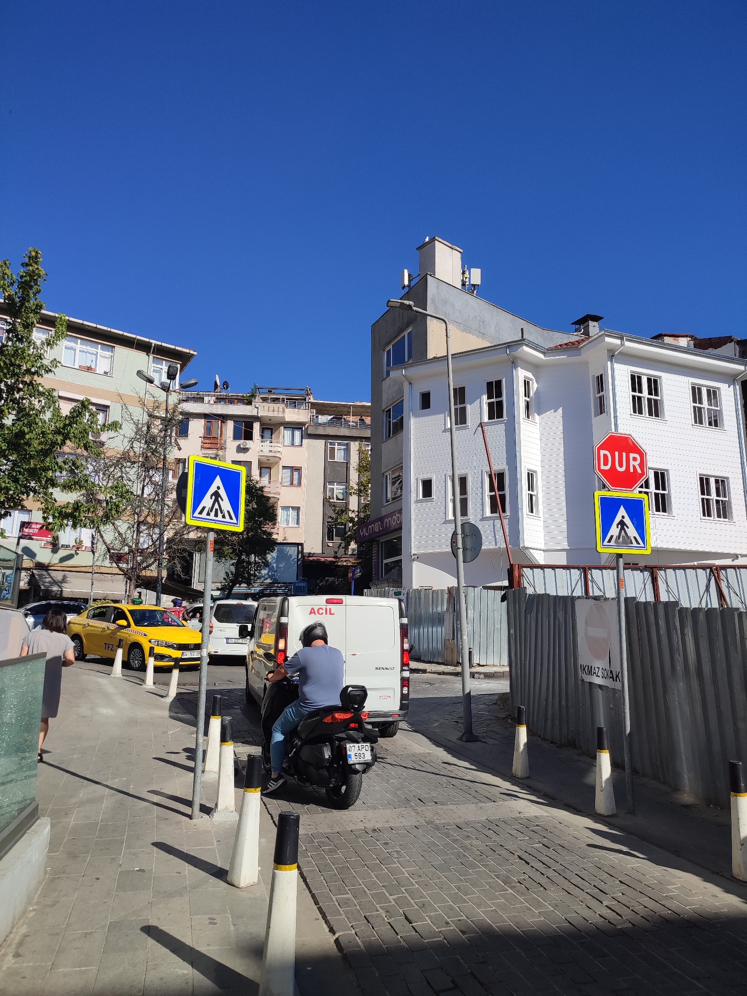 In Üsküdar, Turkey, a sunny day reveals a street scene. A yellow taxi, identified by its license plate "34 TFZ 20," is stopped on the road. Beside it, a white van, bearing the "ACİL" (Emergency) label, is also stationary. A person on a black scooter, wearing a blue shirt and jeans, is in motion, heading away from the viewer. Pedestrian crossing signs are visible. One is a blue square with a white pictogram of a person walking. Another is a red octagonal "DUR" (Stop) sign, with a blue square pedestrian crossing sign below it. A metal fence lines the right side of the scene. Buildings of varying architectural styles and heights, some with visible antennas and satellite dishes, form the backdrop. Trees with green foliage are also present. The sky is a clear, bright blue.