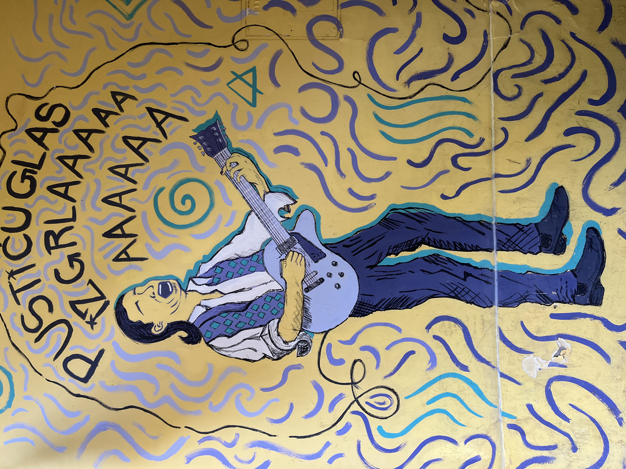 The image shows a section of street art in Novi Sad, Serbia. The artwork depicts a man lying on his back, playing an electric guitar. He is wearing a blue and white patterned shirt and dark blue pants with blue highlights. His mouth is open in what appears to be a scream or song. To the left of the man, the words "PUSTICUGLAS" and "AAAAAAA" are written in black. The background is a yellow-gold color with blue and teal wavy lines suggesting a stylized sound or energy emanating from the musician. A black cable trails from the guitar. A geometric symbol (triangle inside a circle) and a spiral are also present. The artwork is on a wall, which is evident from the visible texture and a patched area in the lower right corner.