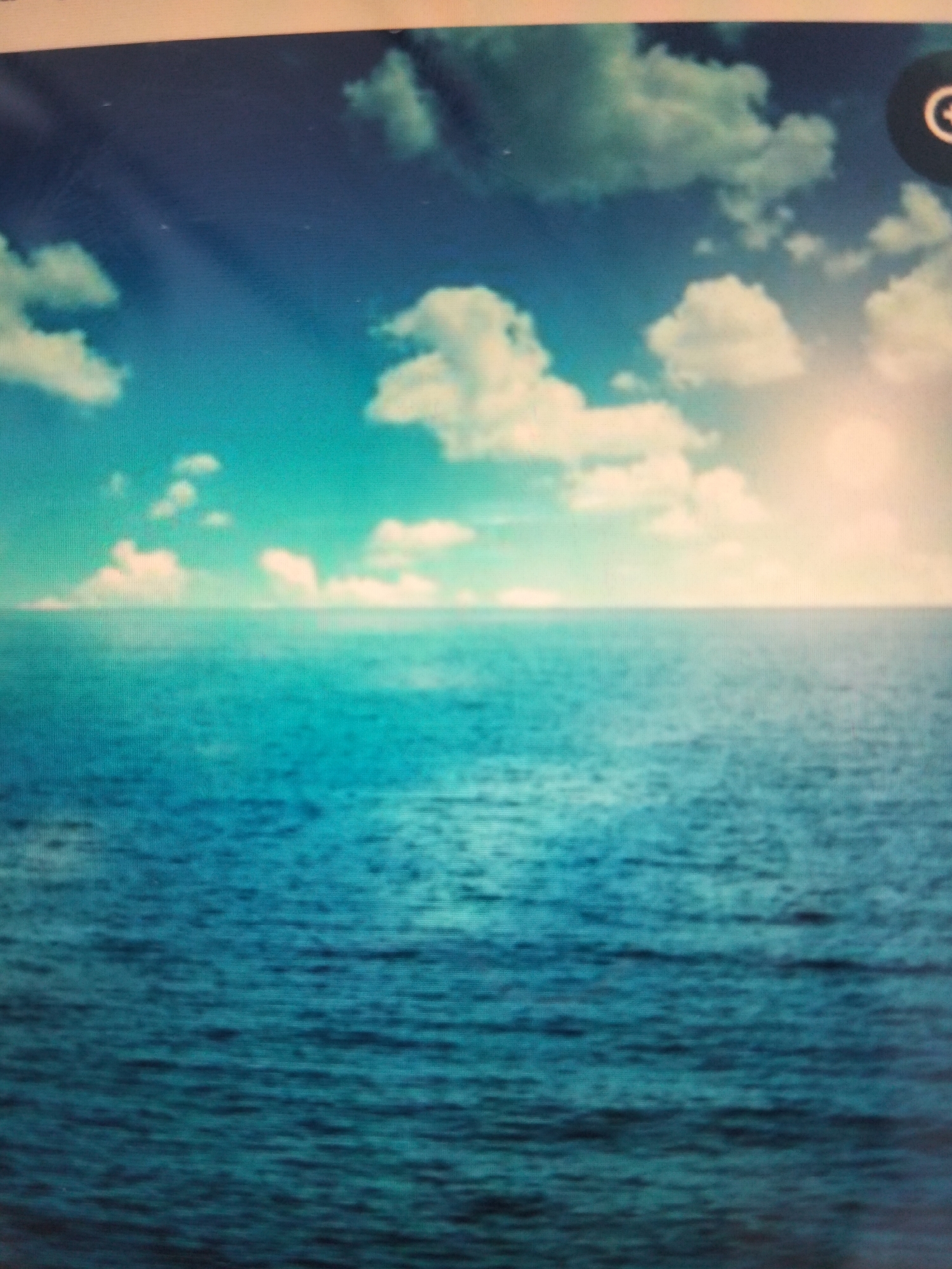 The image depicts a vast expanse of ocean under a bright, partially cloudy sky. The water is a deep blue, with gentle ripples reflecting the light. The horizon is clearly defined, separating the sea from the sky. The sky is a gradient of blues, with fluffy white clouds scattered across it. The sun appears to be setting or rising, casting a warm, golden glow on the clouds and a portion of the ocean. There are no people, buildings, or any other man-made structures visible in the image. The scene conveys a sense of tranquility and natural beauty. Without further information, it is impossible to determine if this scene is located in the city of Payagaboh, Indonesia.