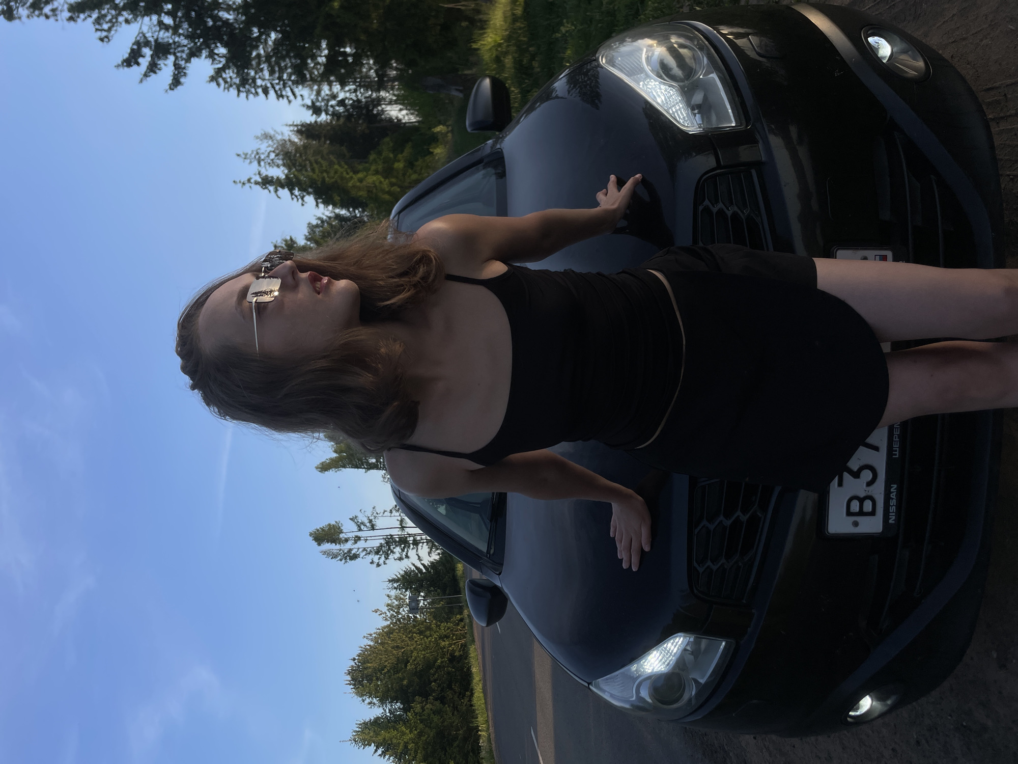 A woman wearing a black top and skirt, and sunglasses, is posing on the hood of a black Nissan car. The car has a license plate that reads "B 37" with the word "NISSAN" printed below the numbers. The car is parked on a paved road, with trees visible in the background. The sky is clear and blue. The photograph appears to have been taken in the Подшивалово region of Russia.