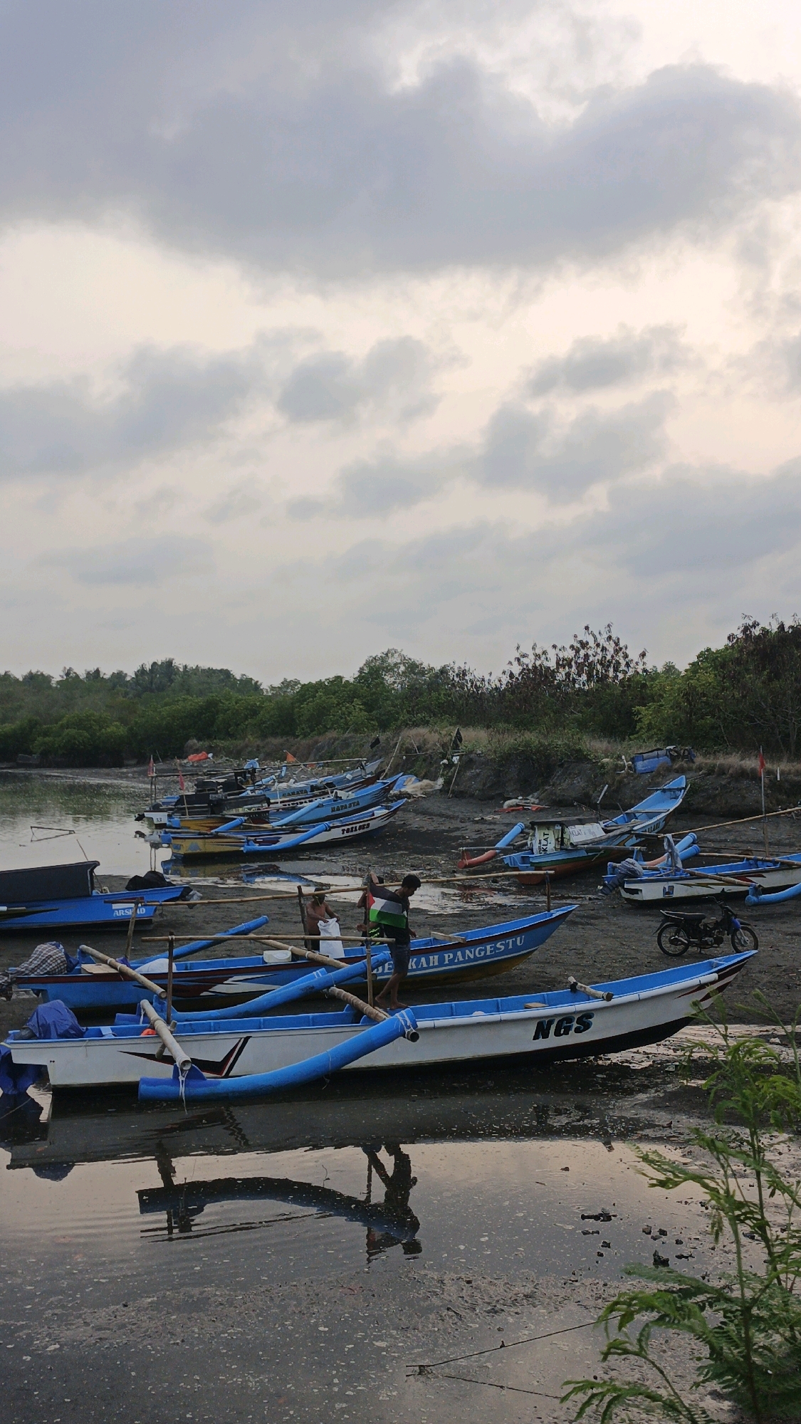 The image depicts a shoreline scene in Desa Karangjaladri, Indonesia, with numerous fishing boats pulled onto the muddy bank. The sky is overcast, with soft, diffused light. In the foreground, several blue and white boats are prominently featured, some partially submerged in the calm, murky water. The boats are of various sizes and appear to be fishing vessels, with some displaying names and numbers such as "NGS" and "KAH PANGESTU". Two men are visible near some of the boats, one appearing to be tending to a fishing net and the other standing. A motorcycle is parked on the bank further back. The background consists of dense green foliage and trees, suggesting a coastal or riverine environment. The overall impression is one of a working fishing village at a quiet time, possibly early morning or late afternoon, given the lighting conditions.