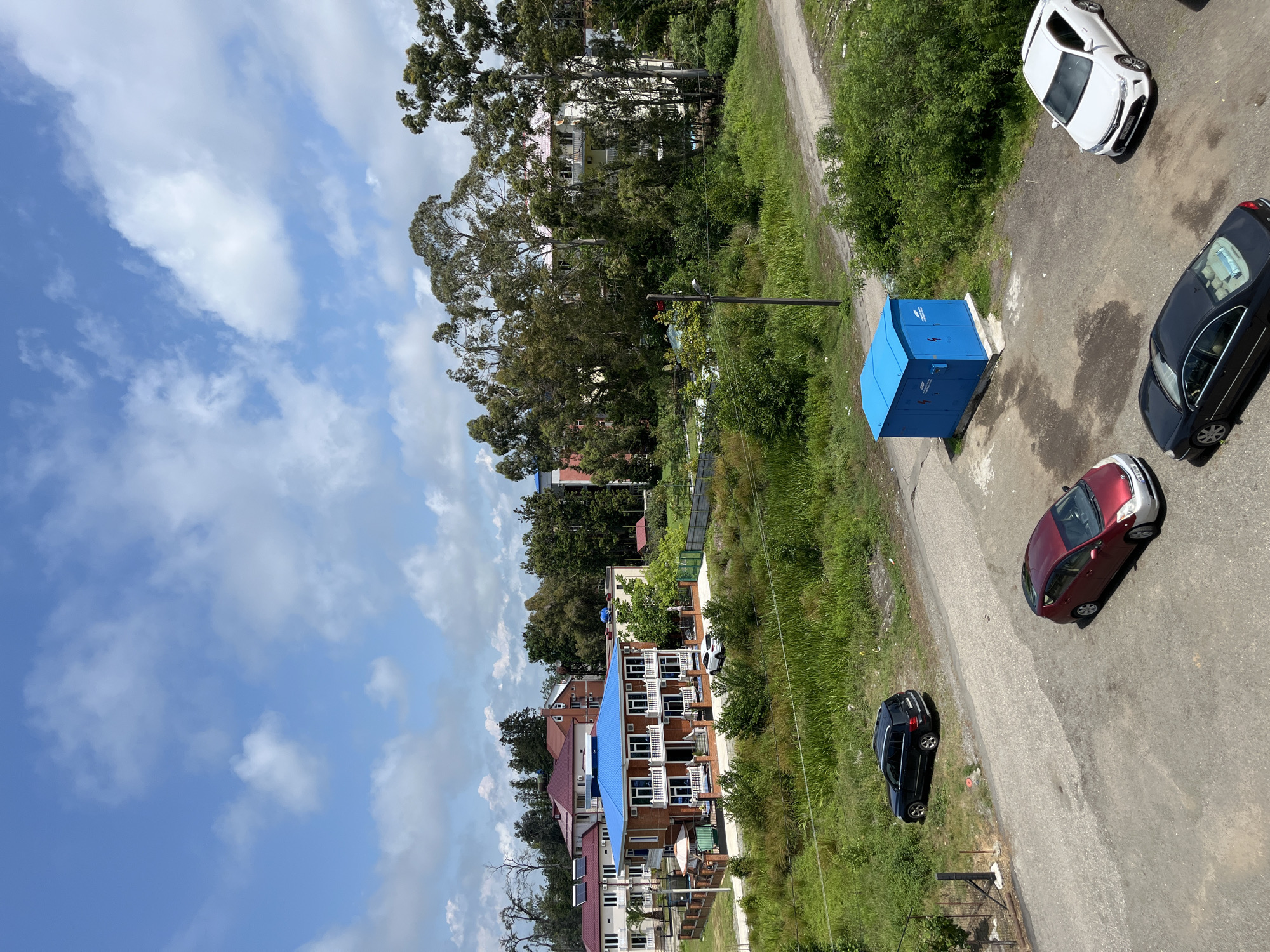 This image depicts an outdoor scene in Ureki, Georgia. The setting appears to be a street or road adjacent to a residential area with multiple buildings and lush greenery. The sky is visible with scattered white clouds against a blue backdrop, suggesting daytime with fair weather.
Several objects are present, including a bright blue electrical transformer box situated on the side of the road. Various cars are parked or driving on the asphalt road: a white car, a black car, and a red and silver car are clearly visible. The buildings in the background vary in architectural style, with some featuring red tiled roofs and others with blue roofs. Some buildings have multiple balconies. Tall trees and dense vegetation line the road, contributing to a natural, somewhat rural or suburban feel.
There are no people clearly visible in the image. The image is taken from an elevated perspective, looking down at an angle. The overall impression is one of a calm, everyday scene in a town or village. No discernible text is visible in the image.