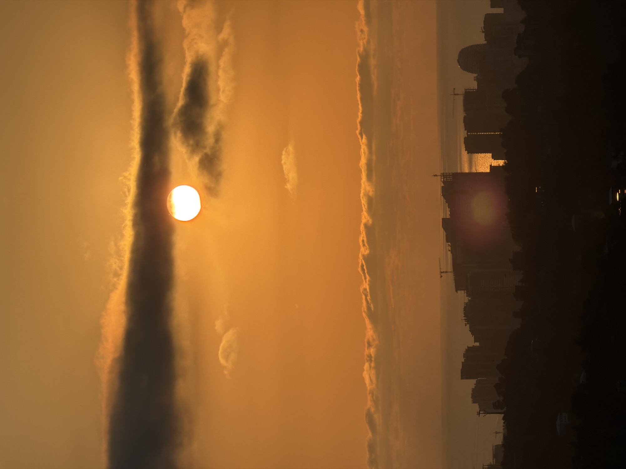 The image captures a sunset over a city, possibly Sameba, Georgia. The sun, a bright orb, is partially obscured by a dark cloud, casting a warm orange glow across the sky. Below, a silhouetted cityscape rises from a dark foreground, with tall buildings and construction cranes visible. The sea can be seen in the distance, reflecting the sunlight. The overall impression is one of a dramatic and atmospheric end to the day.