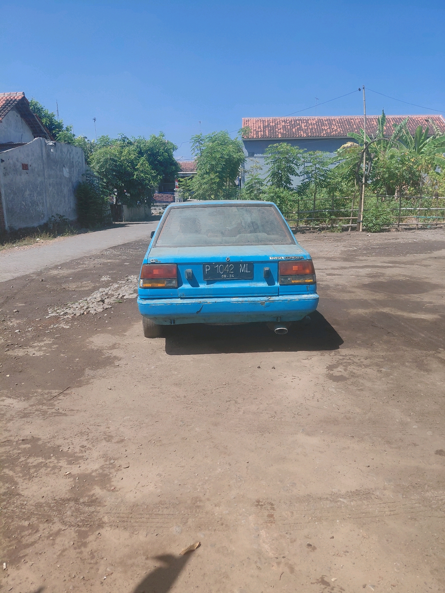 A dusty, light blue Toyota Corolla is parked on a dirt road in Paiton, Indonesia. The car is seen from the rear, with its license plate reading "P 1042 ML". The vehicle appears old and somewhat weathered, with visible dust on its surface and some minor damage to the rear bumper.
The surroundings are rural, with a few buildings visible in the background. One building is a simple concrete structure, while another has a terracotta tiled roof. Lush green trees and foliage are scattered throughout the scene. The sky is a clear, bright blue, suggesting a sunny day. The ground is a mix of dirt, gravel, and some patches of dry grass, indicating an unpaved area. A shadow of a person or object is cast on the ground in the foreground, but the source is not visible in the frame. The overall impression is one of a quiet, undeveloped area under a hot, clear sky.