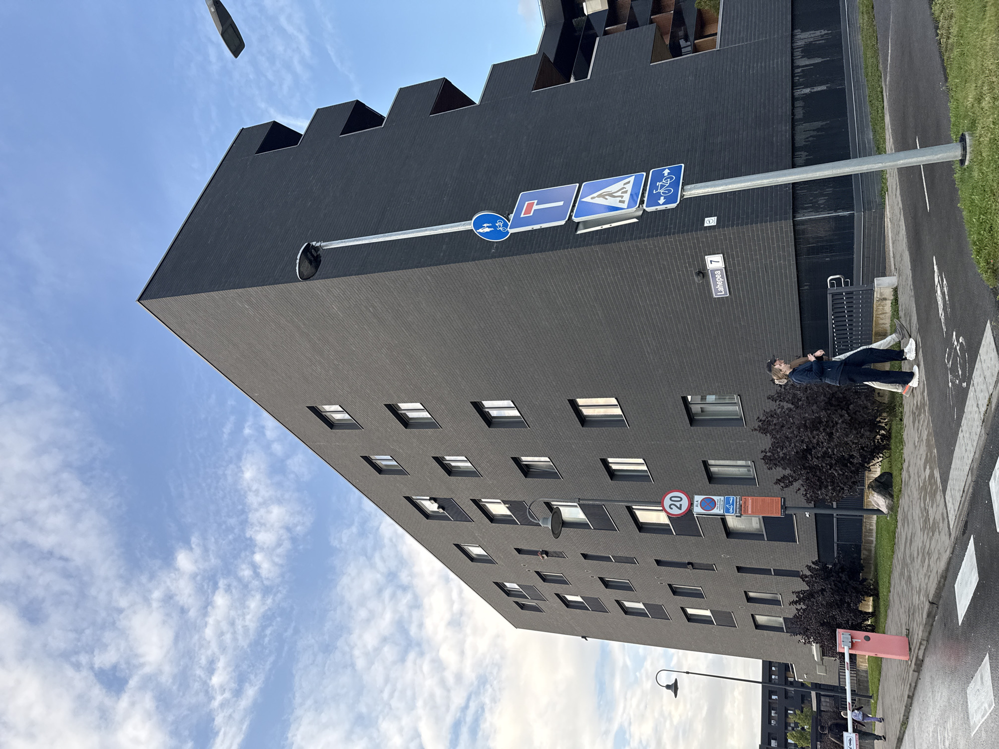 This image depicts a modern, dark gray brick apartment building with a geometrically angular design, located at Lahepea 7 in Tallinn, Estonia. The building features numerous windows and recessed balconies. In the foreground, a sidewalk with a pedestrian crossing is visible. Several street signs are mounted on a pole, including a blue circular sign with a bicycle and pedestrian icon, a red rectangular sign with a white horizontal bar, a blue triangular sign with a pedestrian icon, and a blue rectangular sign with two bicycles and arrows indicating opposite directions. A speed limit sign displaying "20" is also present on a separate pole near the building's entrance. Two women are walking on the sidewalk, one in a dark blue tracksuit and the other in a black jacket and dark pants. A collapsible barrier arm is extended across an entrance to the building's property. The sky is partly cloudy with a clear blue expanse. The overall scene captures a contemporary urban residential environment.