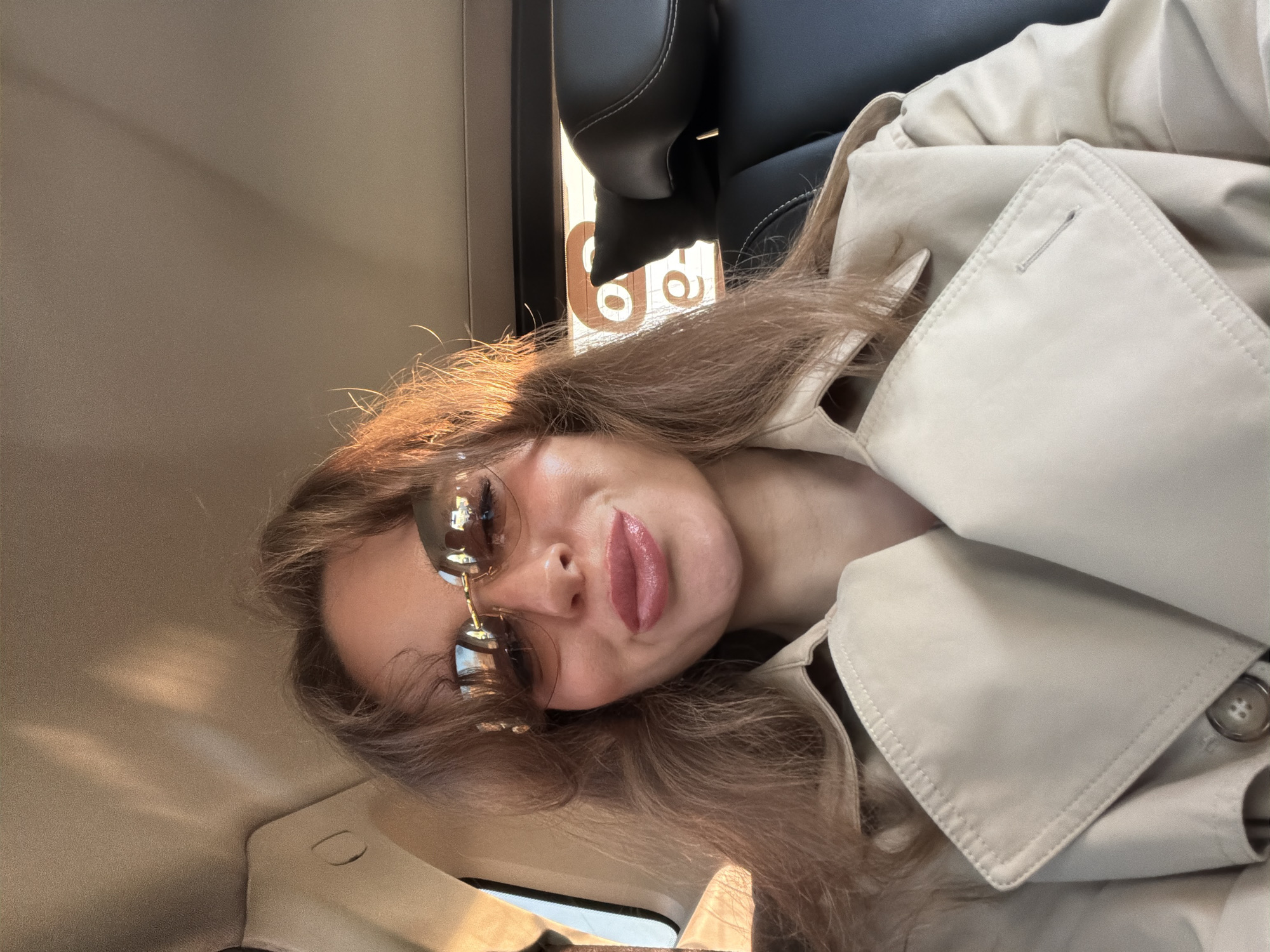 A woman is inside a vehicle, possibly a car or bus. She is wearing a light-colored trench coat and sunglasses. The background shows a glimpse of the vehicle's interior, including a headrest, the side panel, and a window. The lighting suggests it could be daytime. The location cannot be determined from the image alone, nor can the action be described.