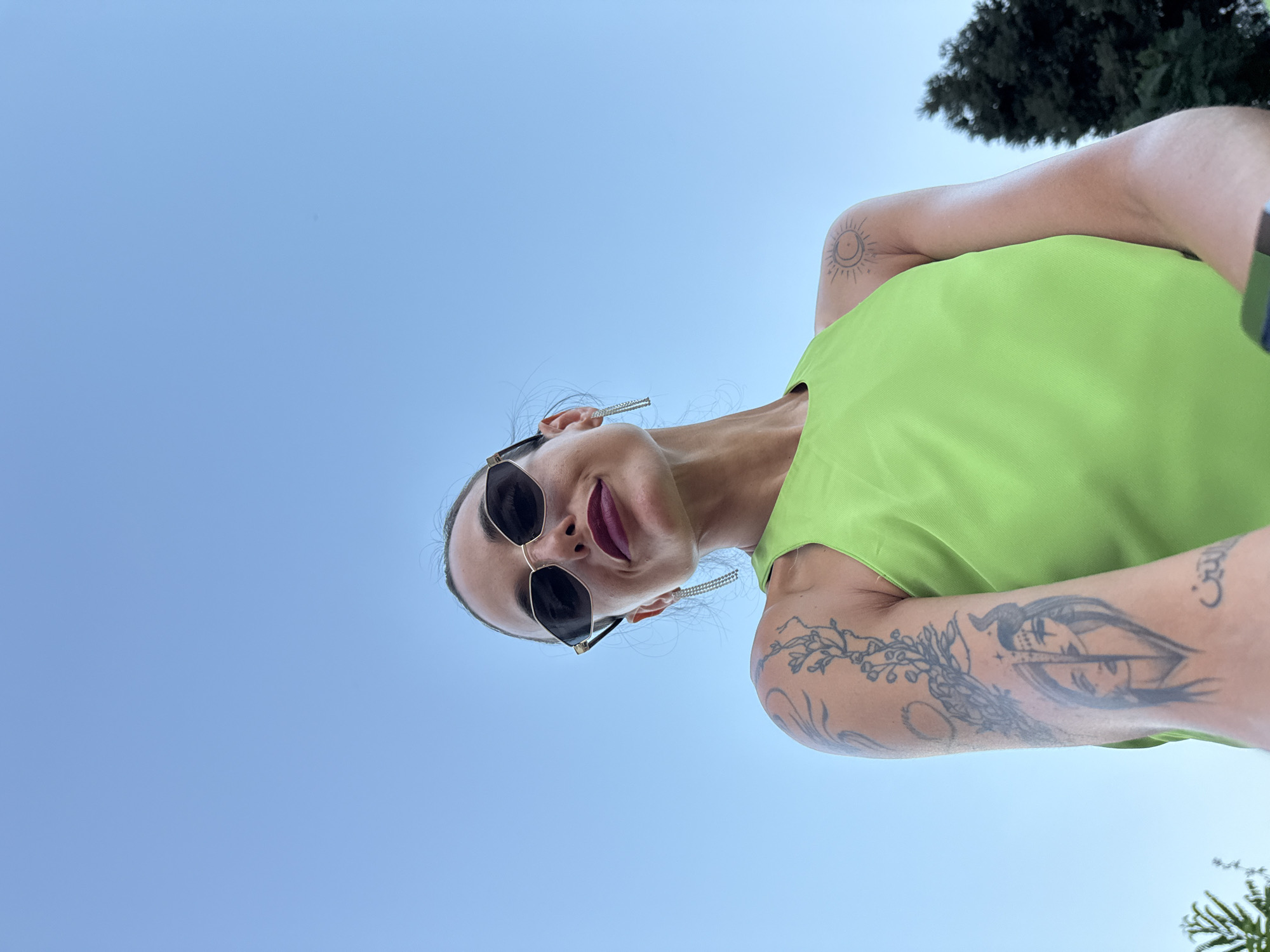 A low-angle, close-up outdoor shot shows a woman with dark hair pulled back wearing oversized sunglasses and a bright green top. She has dark lipstick and long, dangly earrings. Her left arm, visible in the shot, has several tattoos, including one of a face split down the middle and another of a branch with flowers. Her right shoulder also has a tattoo of a sun with a crescent moon. The background is a clear blue sky, with the tops of dark green trees visible in the lower right corner. The image does not provide specific details about the location in Pleven, Bulgaria.