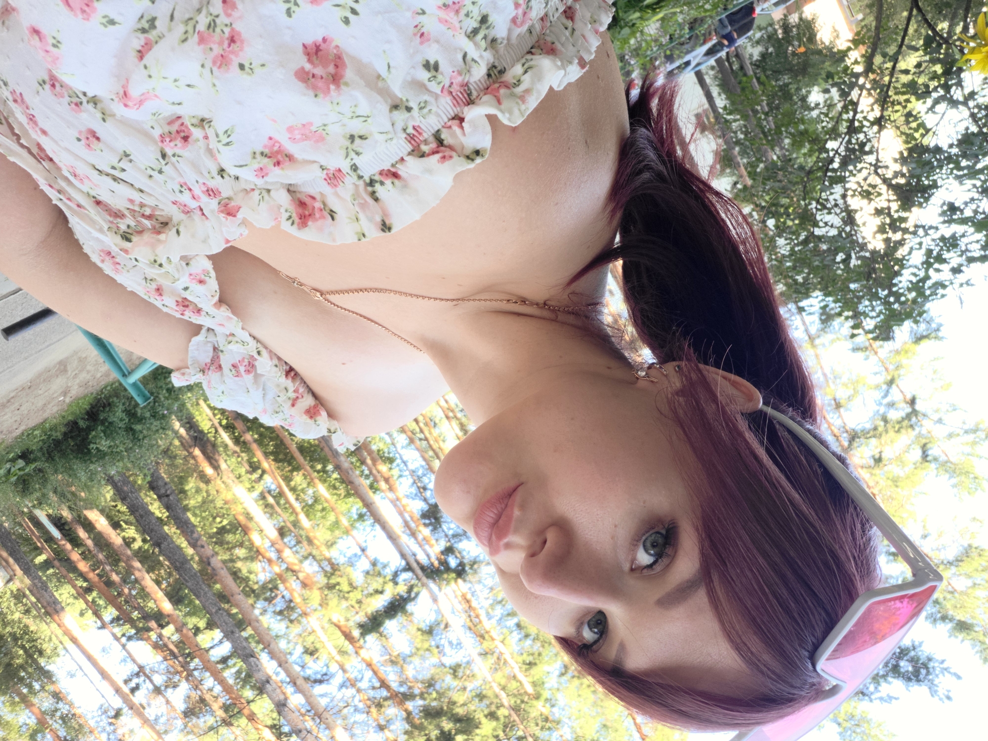 A young woman with auburn hair is photographed from the chest up, outdoors. She is wearing a white off-the-shoulder dress with a pink floral pattern and a delicate gold necklace. Pink-tinted sunglasses are perched on top of her head. Her hair is pulled back into a ponytail, and she is looking directly at the camera with her lips pursed. The background consists of a sun-dappled forest, with tall pine trees and green foliage. The lighting suggests it is daytime. This setting appears to be a park or natural area, possibly within the city of Chaikovsky, Russia, given the commonality of pine forests in the region.