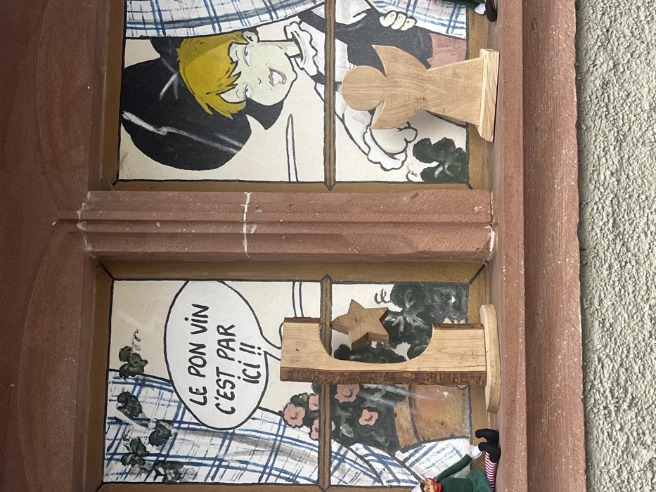 The scene depicts a two-pane window display on a building in Kintzheim, France. The window features reddish-brown stone framing and a central mullion, with a matching stone windowsill below. The wall beneath the sill is light-colored and textured.
Behind the glass, each window pane displays a painted backdrop. The left pane shows a blue and white checkered curtain, a potted plant with pink flowers, and green foliage. A white speech bubble is painted in the upper section, containing the French text "LE BON VIN C'EST PAR ICI !!" (The good wine is here!!). The right pane depicts a cartoon character with bright yellow hair, a smirking expression, a ruffled white collar, and a black hat or garment. A blue and white checkered curtain is partially visible on the far right of this pane, and green foliage is also present. The character's right hand is visible and appears to be pointing towards the left, consistent with the speech bubble's direction.
On the reddish-brown windowsill, several objects are arranged. On the far left, a small elf doll, clad in green and red-and-white striped attire with black shoes, sits facing slightly right. Centered between the panes is a natural wood sculpture with a raw bark edge on one side and a smooth, curved cut-out in its body; a five-pointed wooden star is suspended within this cut-out. To the right of the central mullion, a light-colored wooden silhouette of an angel stands upright on a flat base. A second elf doll, similar to the first, is partially visible on the far right of the windowsill, with only its green leg and black shoe discernible.