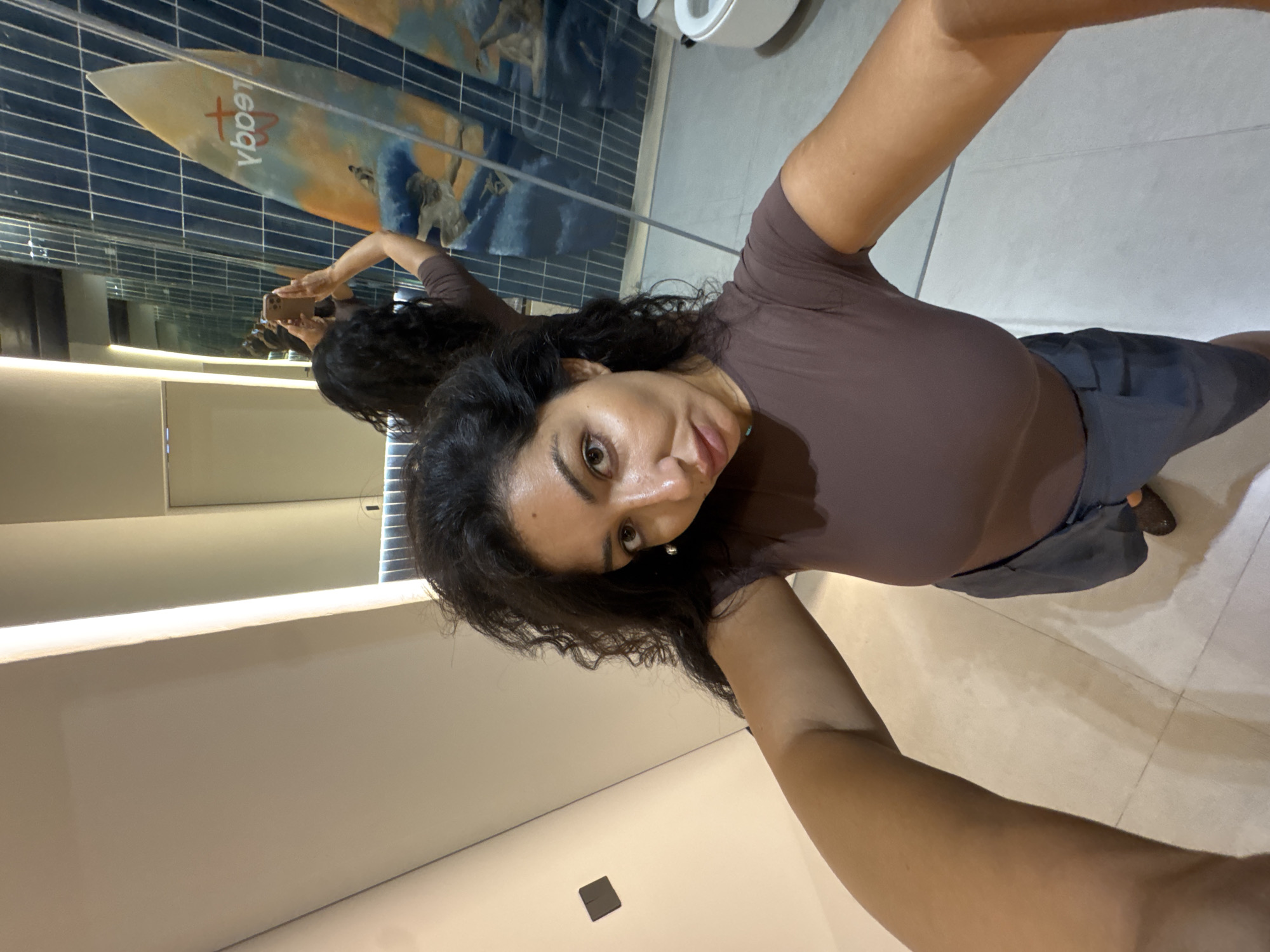 In a mirror selfie taken indoors in Kuta Selatan, Indonesia, a woman with dark, curly hair captures her reflection. She wears a brown form-fitting top and dark shorts or pants. Her face, visible in the mirror, shows a playful expression with wide eyes and a slightly open mouth as she holds a smartphone to take the picture.
Behind her reflection, a distinctive decorative surfboard, featuring the reversed text "legacy" in red, is mounted against a wall adorned with blue rectangular tiles, suggesting a surf-themed or coastal aesthetic. The setting appears to be a modern bathroom or washroom, indicated by the mirror, tiled walls and floor, and a partial view of a white ceramic fixture, possibly a toilet, in the upper right. A linear light fixture illuminates the space below the mirror, and a small dark square object rests on the light-tiled floor. The overall impression is of a casual moment captured in a stylish, well-lit interior.