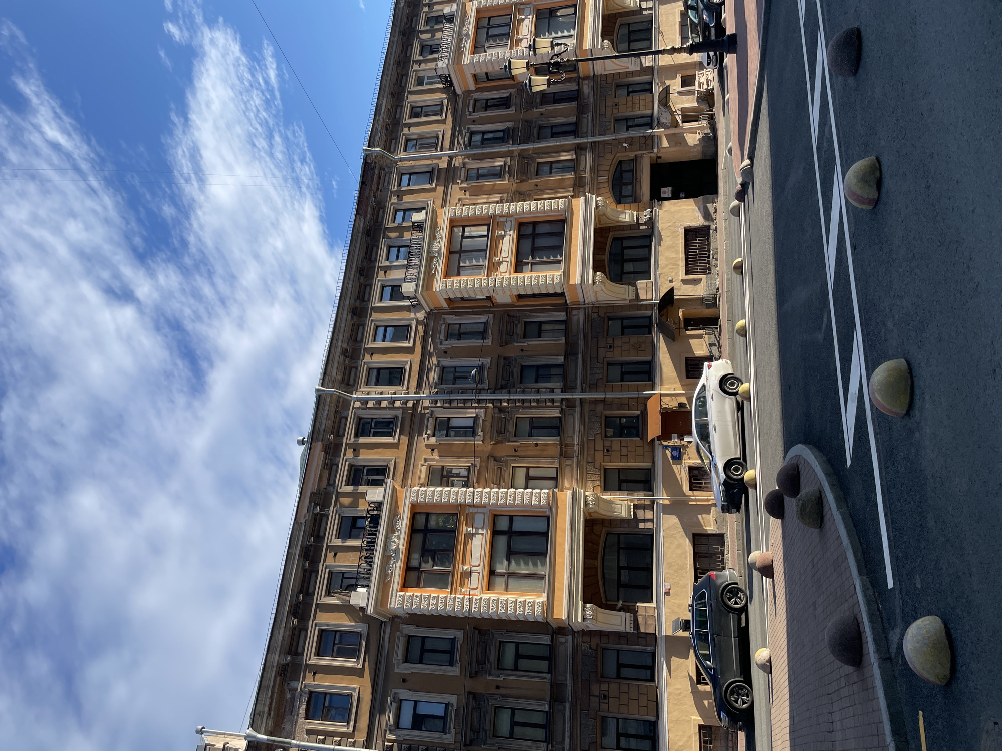 A tall, ornate building is seen under a bright blue sky with scattered clouds. The building, likely located in Saint Petersburg, Russia, has multiple stories with numerous windows, some of which are decorated with elaborate frames. Two cars are parked along the street beside the building. The road is visible with white lane markings and round stone-like objects lining the edge of the sidewalk. Overhead wires stretch across the sky.