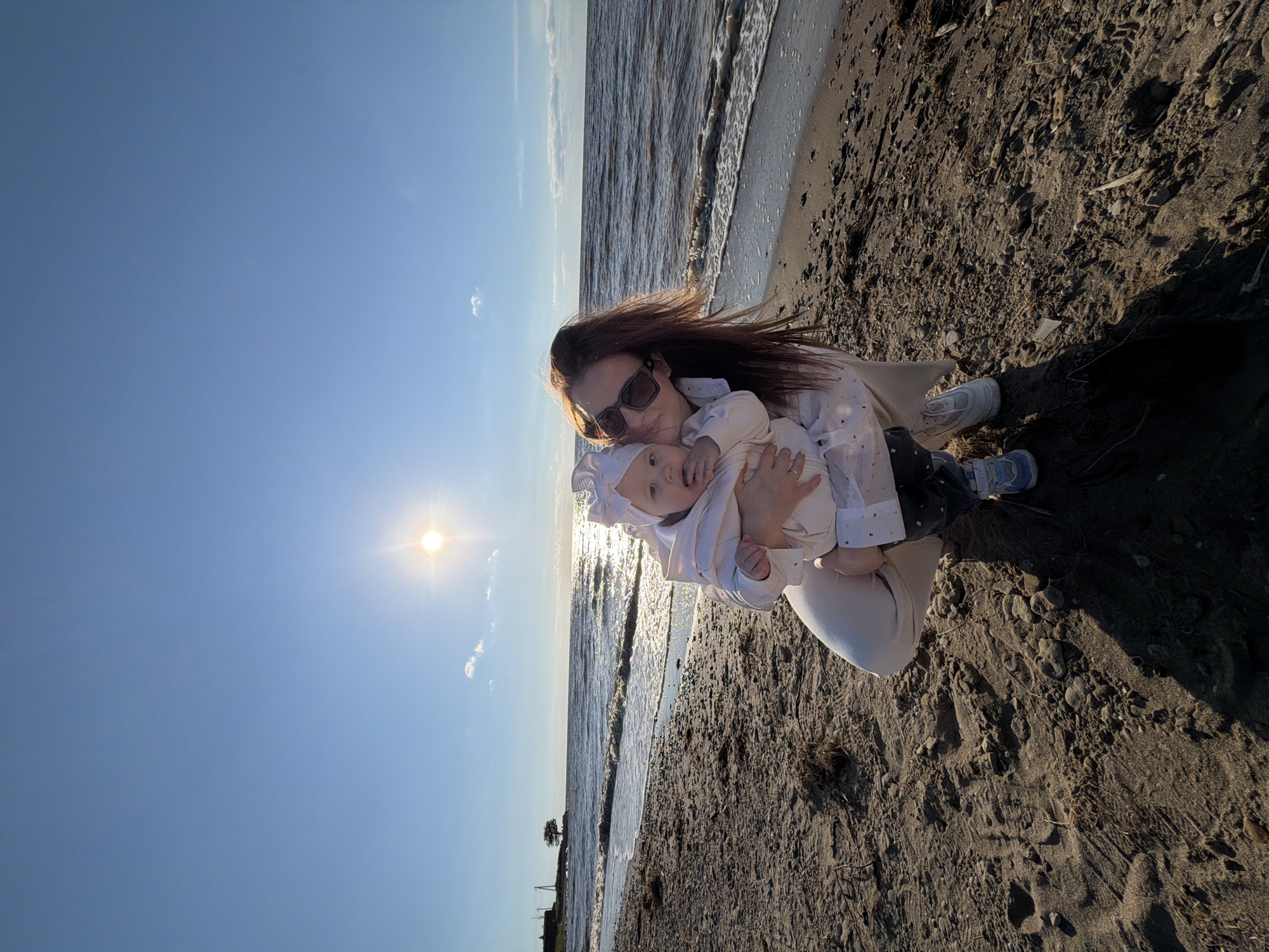 In Port-Katon, Russia, a woman kneels on a sandy beach, holding a baby in her arms. The sun is low in the sky, casting a warm glow over the scene. The sea stretches out behind them, with gentle waves lapping at the shore. A single tree stands on the horizon, silhouetted against the bright sky. The woman is wearing sunglasses and a light-colored outfit, while the baby is dressed in a white outfit and bonnet. The beach is covered in sand and small stones, with some dried seaweed scattered about. The overall atmosphere is one of peacefulness and natural beauty.