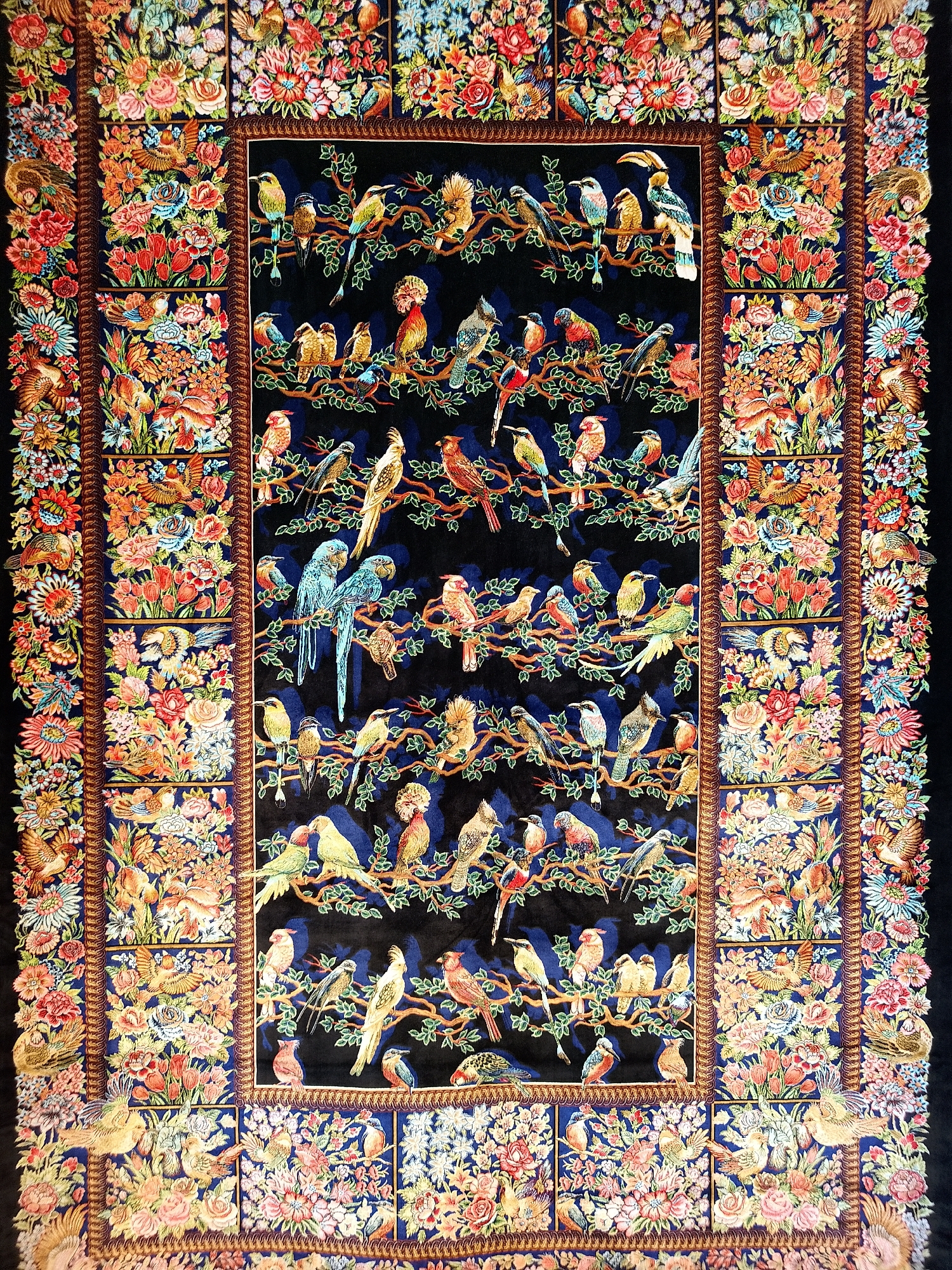 The image shows a richly detailed tapestry or rug, predominantly black in the central field. This field features numerous colorful birds perched on branches against the dark backdrop. The border of the tapestry is densely populated with floral motifs and stylized birds arranged in a grid-like pattern. The style suggests an oriental or Persian aesthetic. The location is Unknown, Unknown.