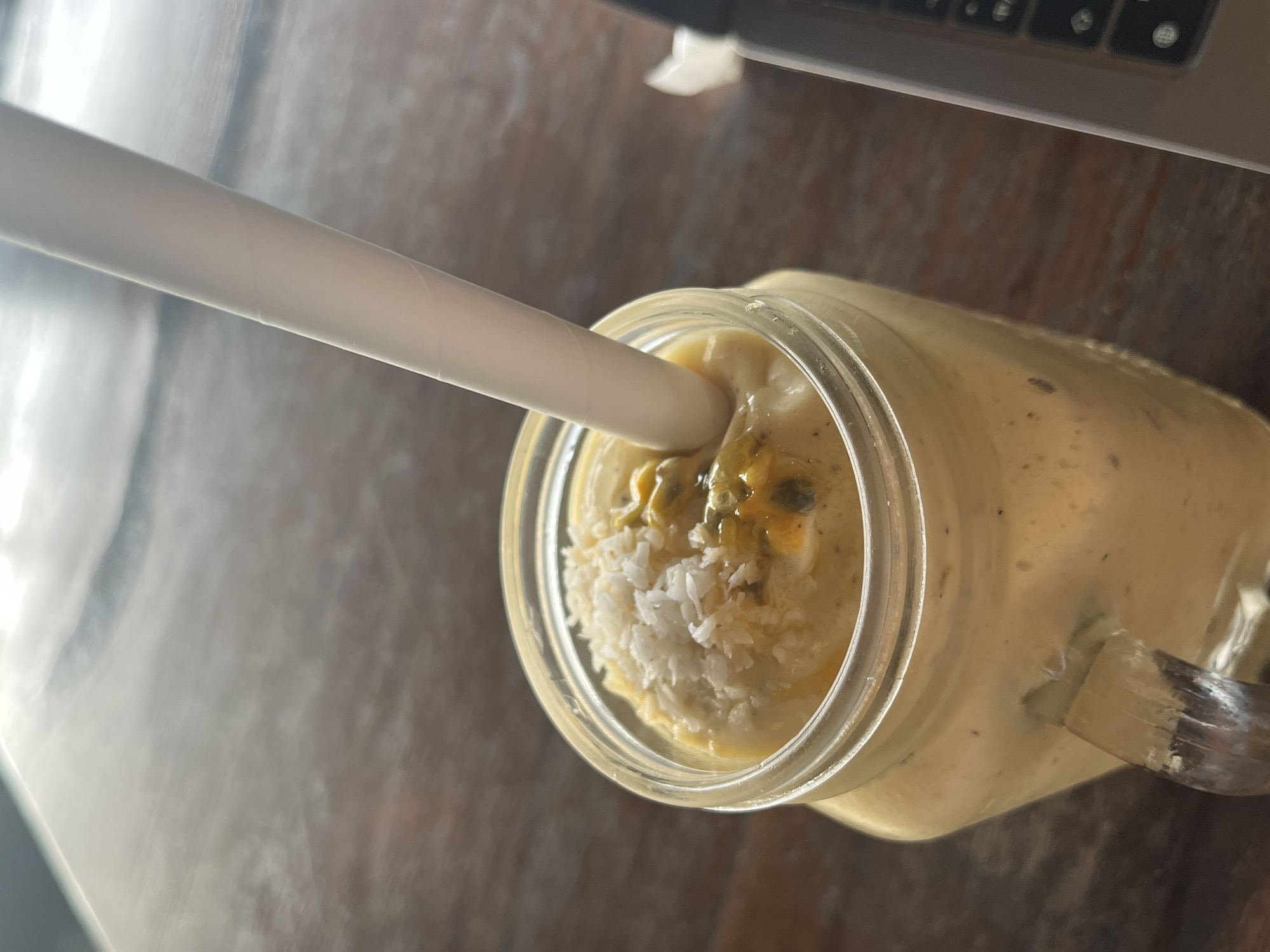 A close-up, overhead shot captures a clear glass mason jar filled with a light yellow smoothie. A white paper straw is inserted into the smoothie, with its end submerged in the liquid. The smoothie is topped with shredded coconut and visible passion fruit seeds and pulp. The jar has a metal handle, and the contents suggest a refreshing beverage. The setting appears to be a table with a textured, dark wood grain, likely indoors given the artificial lighting and the presence of a laptop keyboard at the top right of the frame. The overall ambiance is casual and relaxed, possibly indicating a moment of personal enjoyment or work-life balance. The context provided, Baan Chaweng Noi, Thailand, hints at a tropical location.
