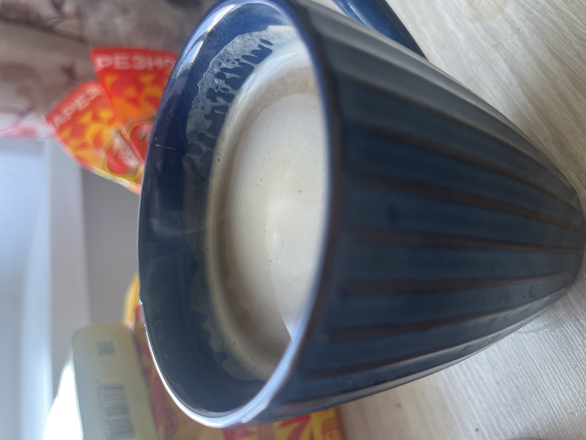 A close-up, eye-level shot shows a blue ceramic mug filled with a frothy, light brown beverage, likely coffee or latte. The mug has vertical, ridged texturing. Wisps of steam rise from the surface of the drink. In the background, slightly out of focus, are two items: on the left, a light yellow rectangular box with black text, and on the right, a partially obscured bag with bright orange and red packaging that has some lettering visible. The objects are placed on a light-colored, textured surface, possibly a wooden table. The overall lighting suggests an indoor setting with natural light coming from the left. There are no people or animals present in the image. The scene is static. The image does not provide any information about the city of Chelyabinsk, Russia.