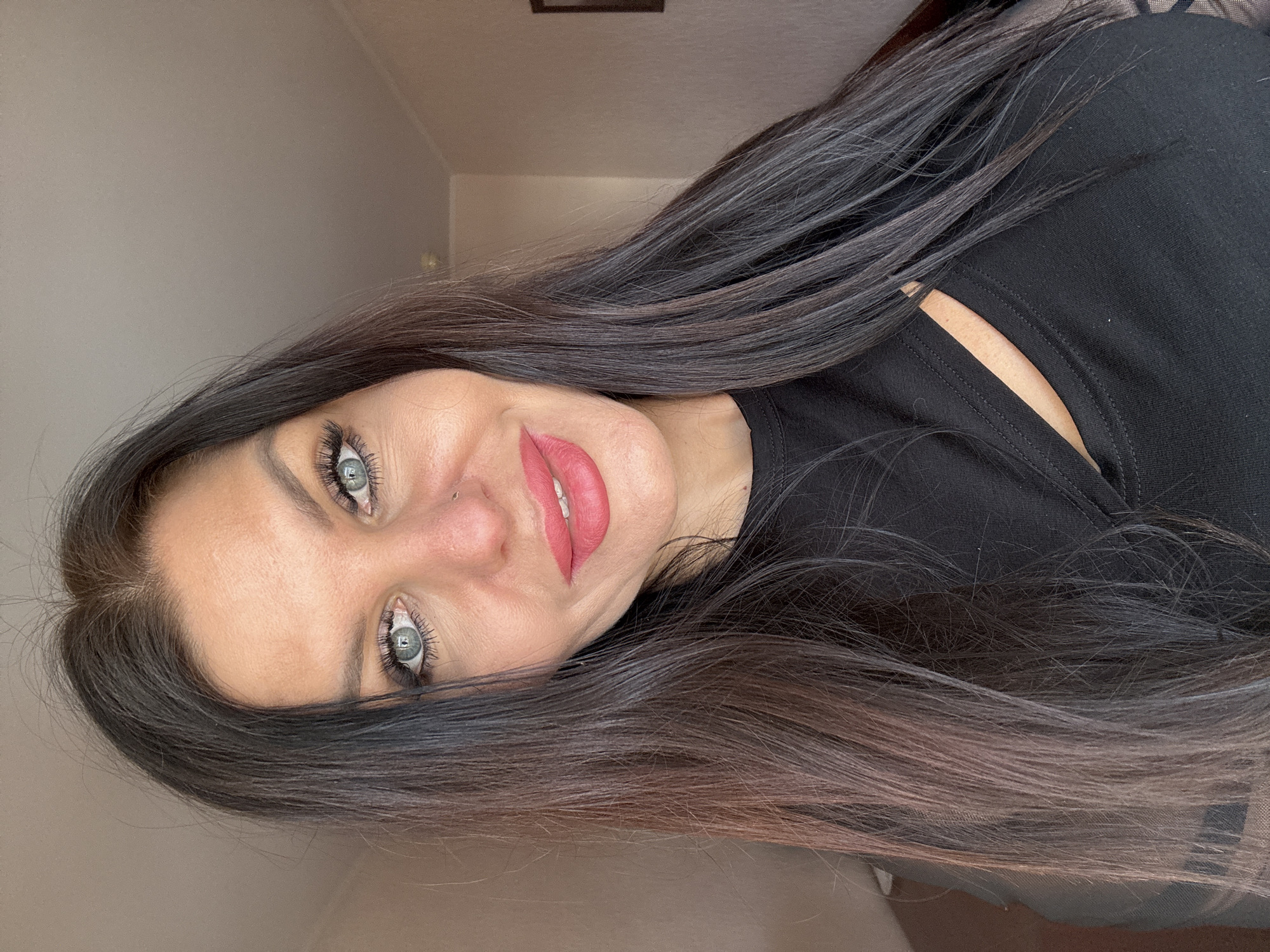 A head-and-shoulders portrait depicts a woman from Perm, Russia, looking directly at the camera with a gentle, engaging smile. Her long, dark hair, with subtle reddish-brown highlights towards the ends, frames her face and cascades down her shoulders. She has striking light-colored eyes, accentuated by prominent dark mascara and eyeliner, and well-defined eyebrows. A small stud piercing is visible on her left nostril, and her lips are colored with a matte or satin reddish-pink lipstick. She wears a plain black top or dress, which features a small cut-out detail revealing a sliver of skin near her right shoulder/collarbone area. The background is a softly blurred, light-colored indoor wall, with a faint rectangular object (possibly a picture frame) visible in the upper portion. The lighting is soft and even, suggesting an indoor setting, without indicating a specific time of day or weather. No text is visible in the image.