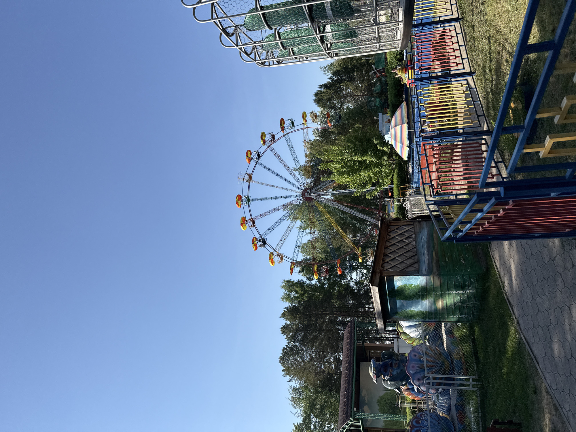 The image shows a Ferris wheel in an amusement park setting, captured on a clear, sunny day under a bright blue sky. The Ferris wheel, featuring carriages in shades of yellow and orange, is positioned amidst lush greenery, suggesting a park or recreational area. The lower portion of the image reveals elements of other amusement park rides or attractions, including colorful structures and enclosed areas with fencing, hinting at a variety of activities available in this recreational space. The location is in the city of Unknown, Unknown.