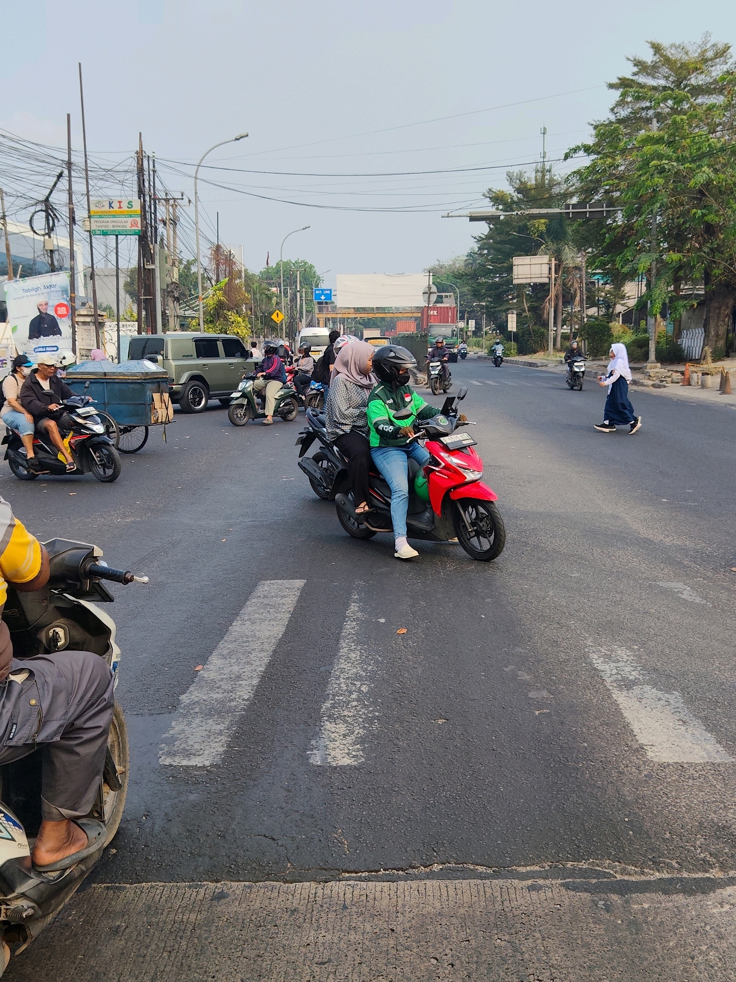 The image depicts a street scene in Bekasi, Indonesia, likely in a busy urban area. Several motorcycles, some with multiple passengers, are visible on the asphalt road. On the right side of the road, a young person in a white hijab and dark blue skirt is walking across a pedestrian crossing. In the foreground, a person on a motorcycle is partially visible from the waist down, wearing gray pants and a sandal.
The environment includes utility poles with a dense network of electrical wires, streetlights, trees lining the roadside, and buildings in the background. One building displays a sign that reads "KIS" with a logo, and another banner features an image of a man and text in Indonesian. Traffic signs, including a "BUS LINE" sign, are also present. The road has visible white pedestrian crossing lines. The overall impression is that of a typical day on a moderately busy street in an Indonesian city.