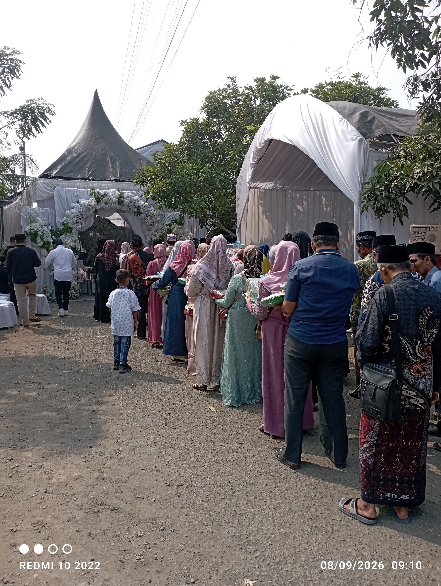 In Dukuhseti, Indonesia, an outdoor event is taking place under a large white tent and a conical tent. A line of people, predominantly women in modest Islamic attire, are gathered, holding trays of food. Men in similar traditional clothing are also present. A young boy in a patterned shirt stands facing away from the camera. The ground is a dusty, gravelly surface. Decorative white floral arrangements adorn the entrance to the white tent. The scene suggests a communal gathering or celebration, possibly a wedding reception or religious festival.