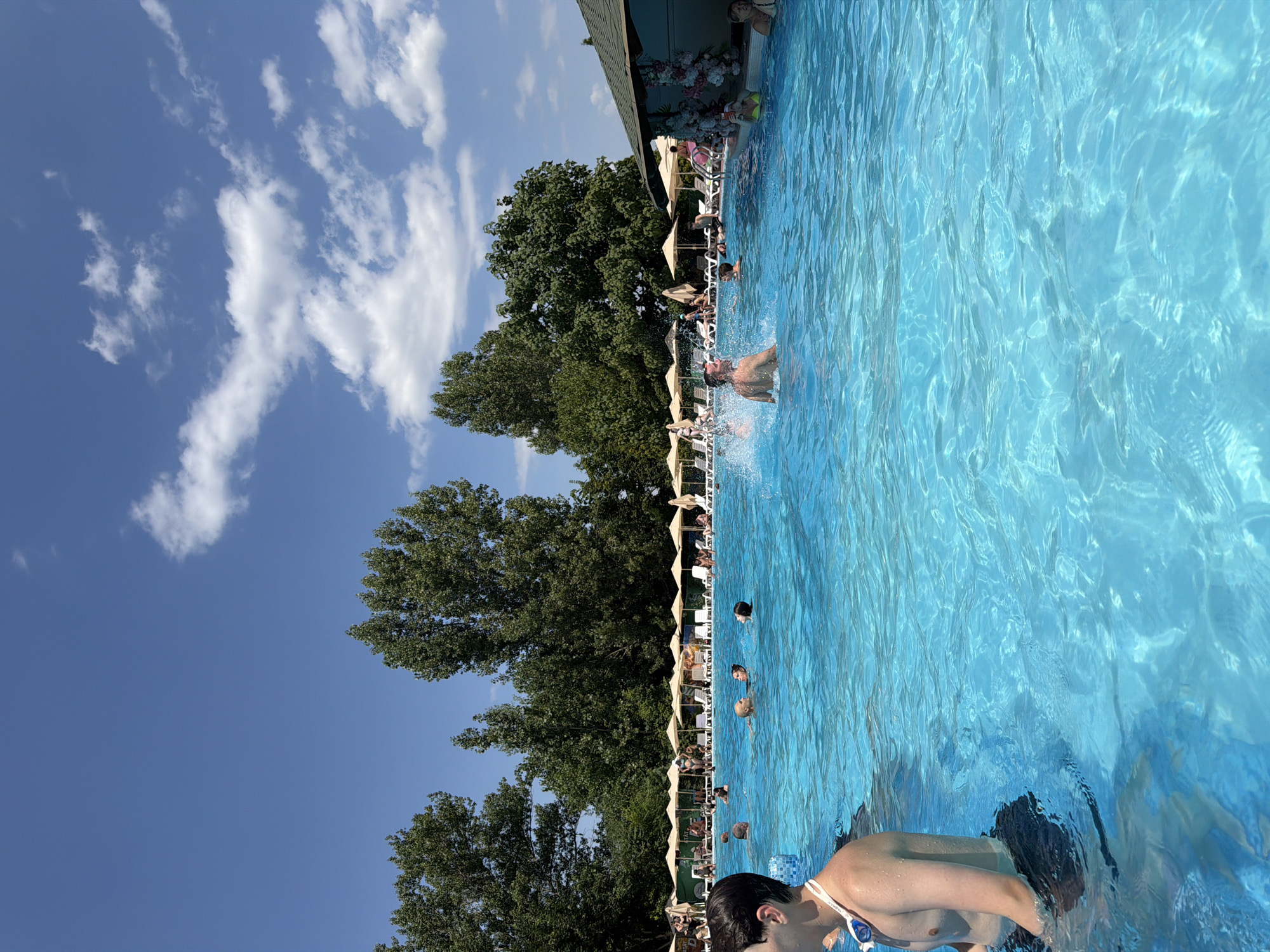 The image depicts a crowded outdoor swimming pool on a sunny day, likely at the "Curortul pentru odihna si sanatate" in Sociteni, Moldova, as indicated by the context provided. The pool is filled with clear blue water, and several people are engaged in various activities. In the foreground, a young man with dark hair and goggles around his neck stands partially submerged in the water, facing away from the camera. In the mid-ground, a man is splashing water, creating a noticeable disturbance on the surface. Other individuals are seen swimming, talking, and lounging by the poolside.
The background reveals a row of white lounge chairs topped with umbrellas, providing shade for pool-goers. Beyond the pool area, a dense line of mature green trees forms a natural backdrop. A small, roofed structure, possibly a bar or service area, is visible to the right, adorned with floral decorations. The sky above is a vibrant blue, dotted with fluffy white clouds, suggesting fair weather. The overall scene captures a lively atmosphere of recreation and relaxation at a public pool.