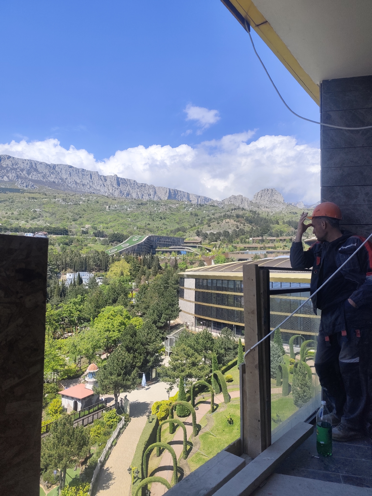 From a high vantage point, the image captures a broad view of Алупкинский городской совет, Ukraine. Prominently featured is a worker wearing an orange hard hat and dark clothing, standing on what appears to be a construction site or balcony. He is positioned to the right of the frame, with his hand raised to his head as if shading his eyes or in contemplation. Another person is partially visible beside him. The foreground includes building elements under construction. The midground reveals a lush landscape with manicured gardens, pathways, and buildings. A distinctive architectural structure with a green roof can be seen further back, among trees. In the background, a mountain range dominates the skyline under a partly cloudy sky.
