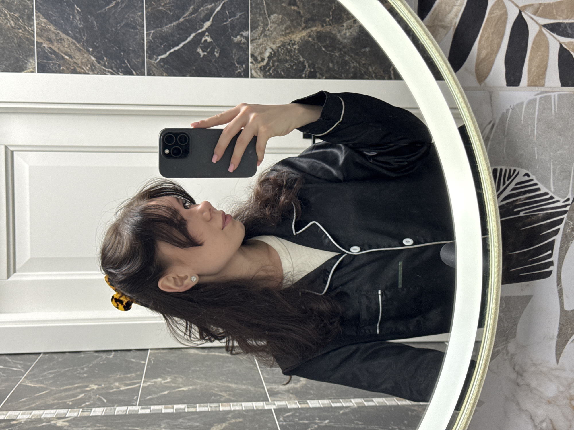 A woman in a black satin pajama top is taking a selfie in a bathroom. The reflection in the mirror shows her holding a smartphone. The bathroom features a dark-tiled floor and marble-patterned tiles on the wall, contrasted with a white paneled wall. A round mirror with an integrated light illuminates the scene. The location is Unknown, Unknown.