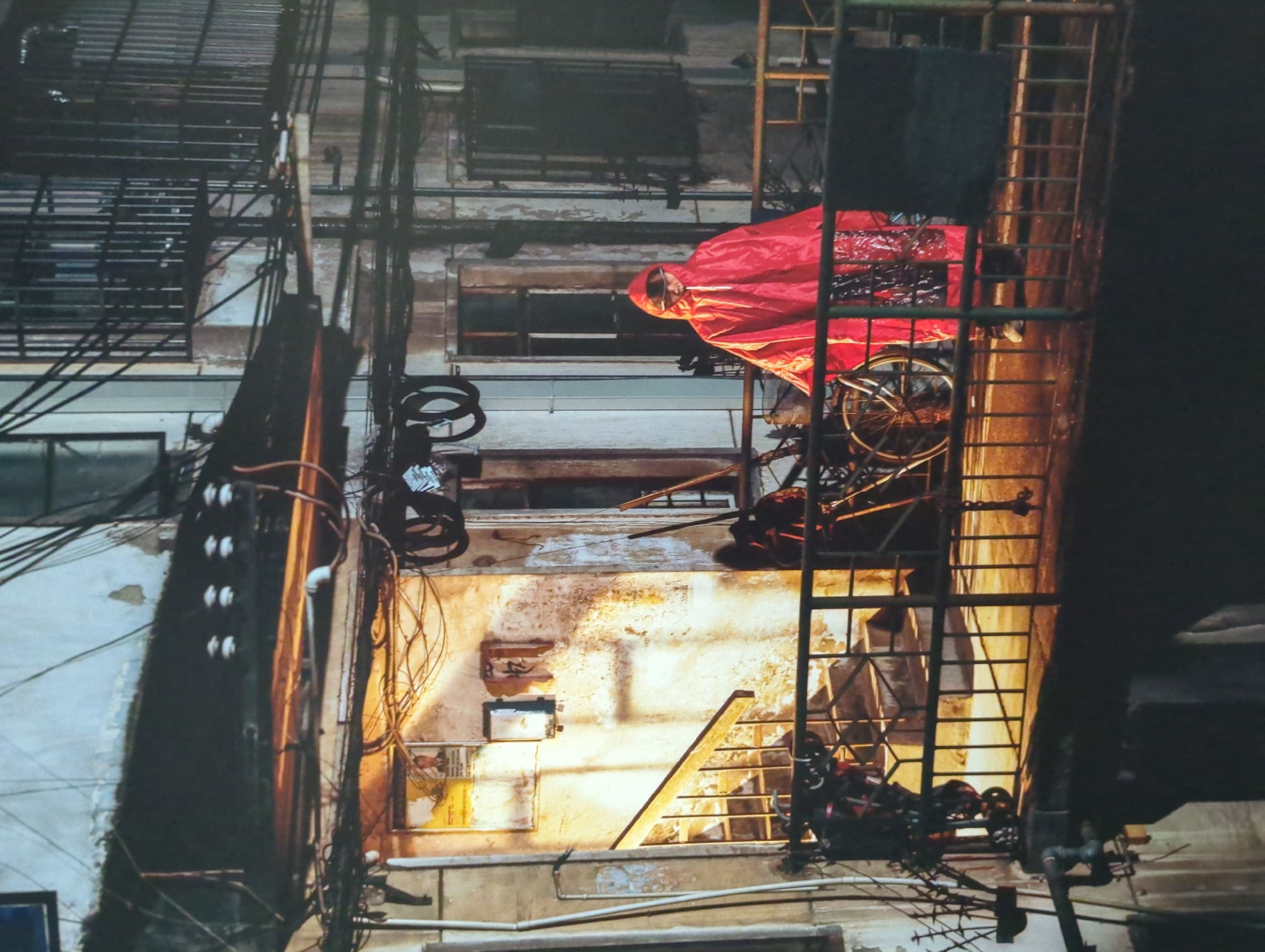 A person wearing a red hooded raincoat stands on a metal balcony in what appears to be an urban residential area in Jinniu District, China. The person is facing the camera, with their hands inside the coat. A bicycle is parked behind them on the balcony. The balcony overlooks a dimly lit outdoor staircase. The building walls are weathered and show signs of peeling paint. Numerous electrical wires and conduits are visible crisscrossing the exterior of the buildings. The scene is lit by a combination of warm artificial light and the ambient darkness of the evening.